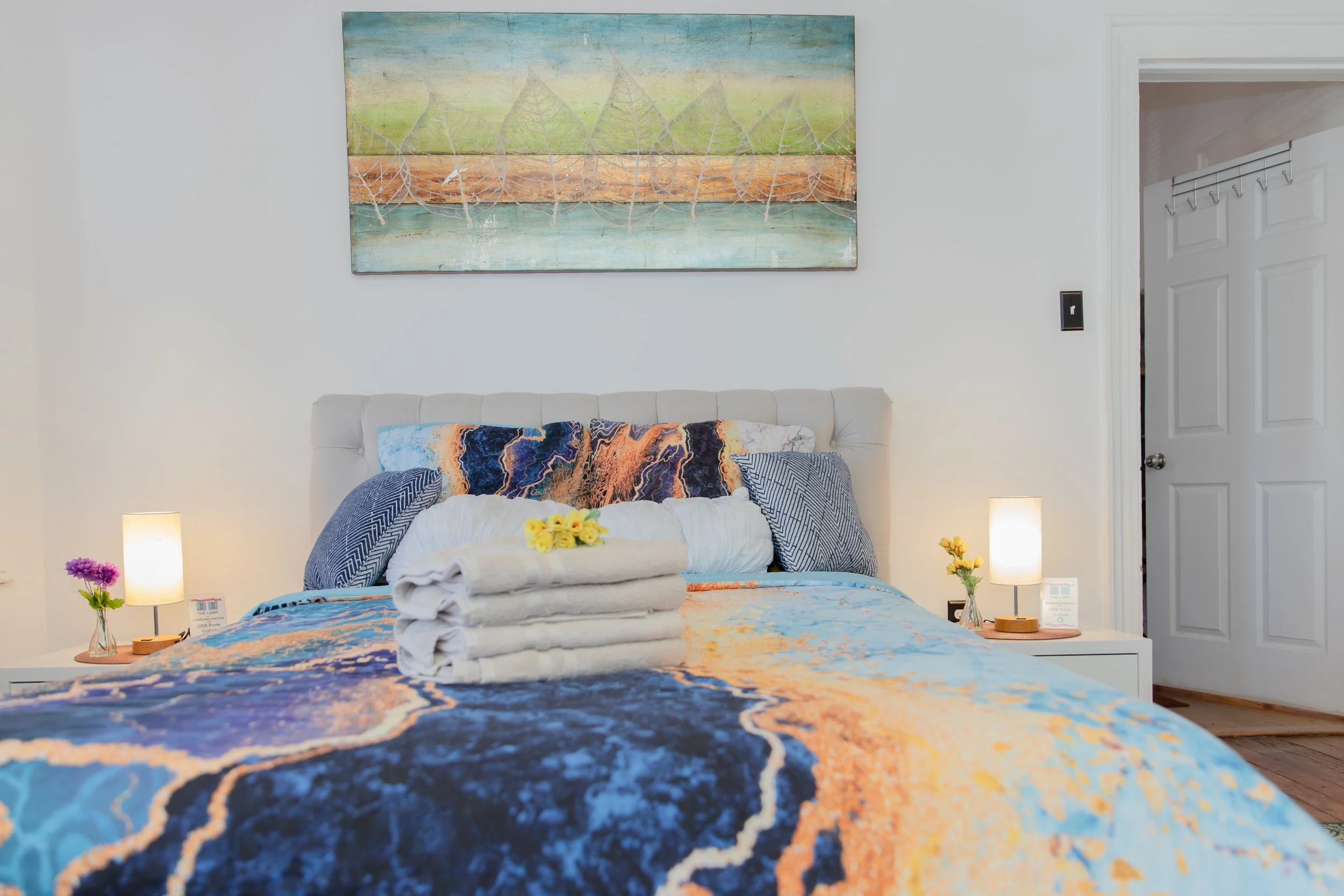 A neatly made bed with a colorful abstract bedspread in shades of blue, orange, and yellow, topped with rolled white towels and a small flower. The headboard is white and tufted, with two patterned blue pillows and a decorative pillow with vibrant orange, navy, and white patterns. There are small bedside tables on each side with lamps and flowers, and a large abstract landscape painting hangs above the bed. A doorway is visible on the right.