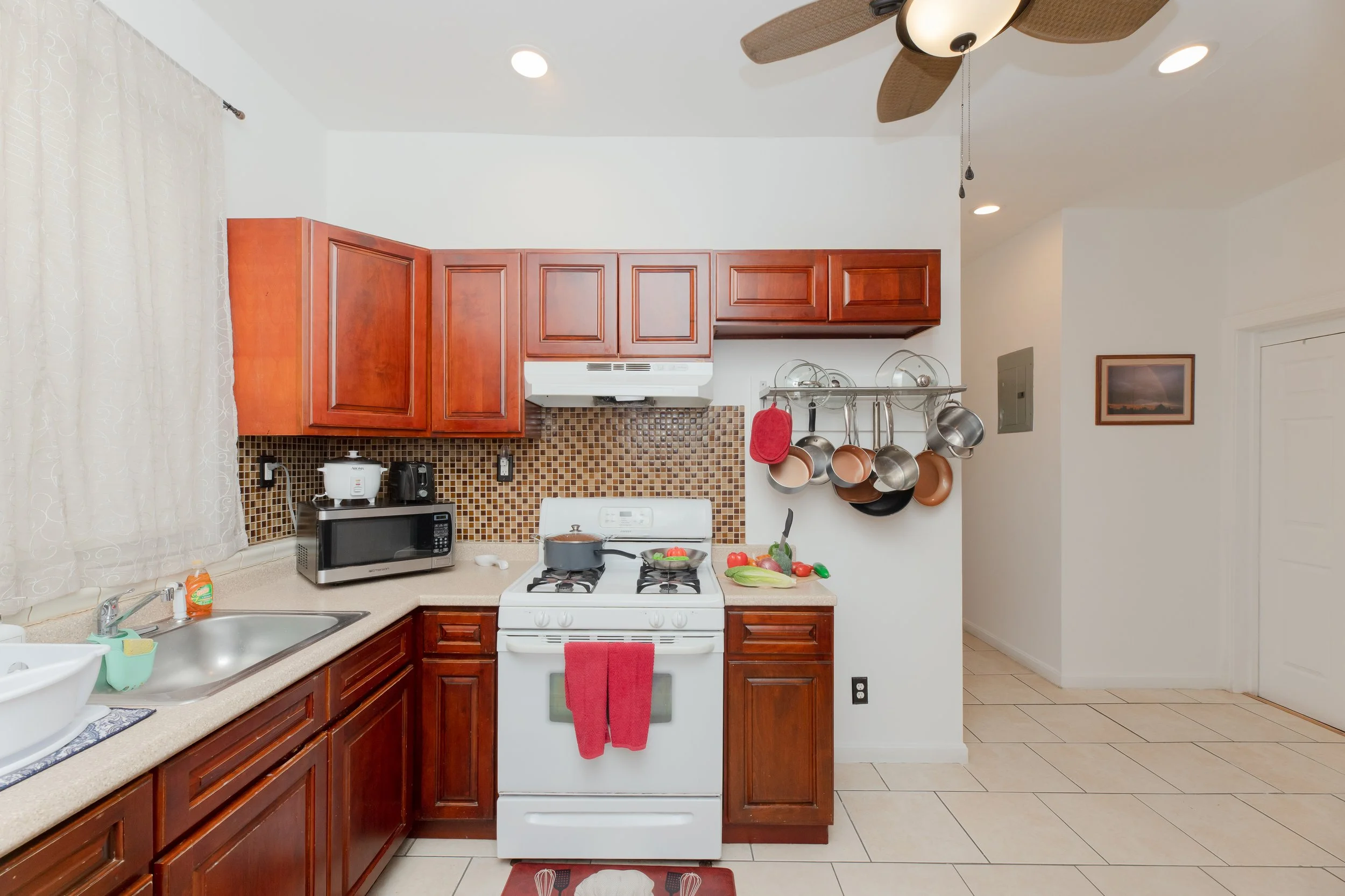 Kitchen with white stove, wooden cabinets, microwave, sink, and hanging pots and pans, alongside a window with curtains and beige tiled floor.