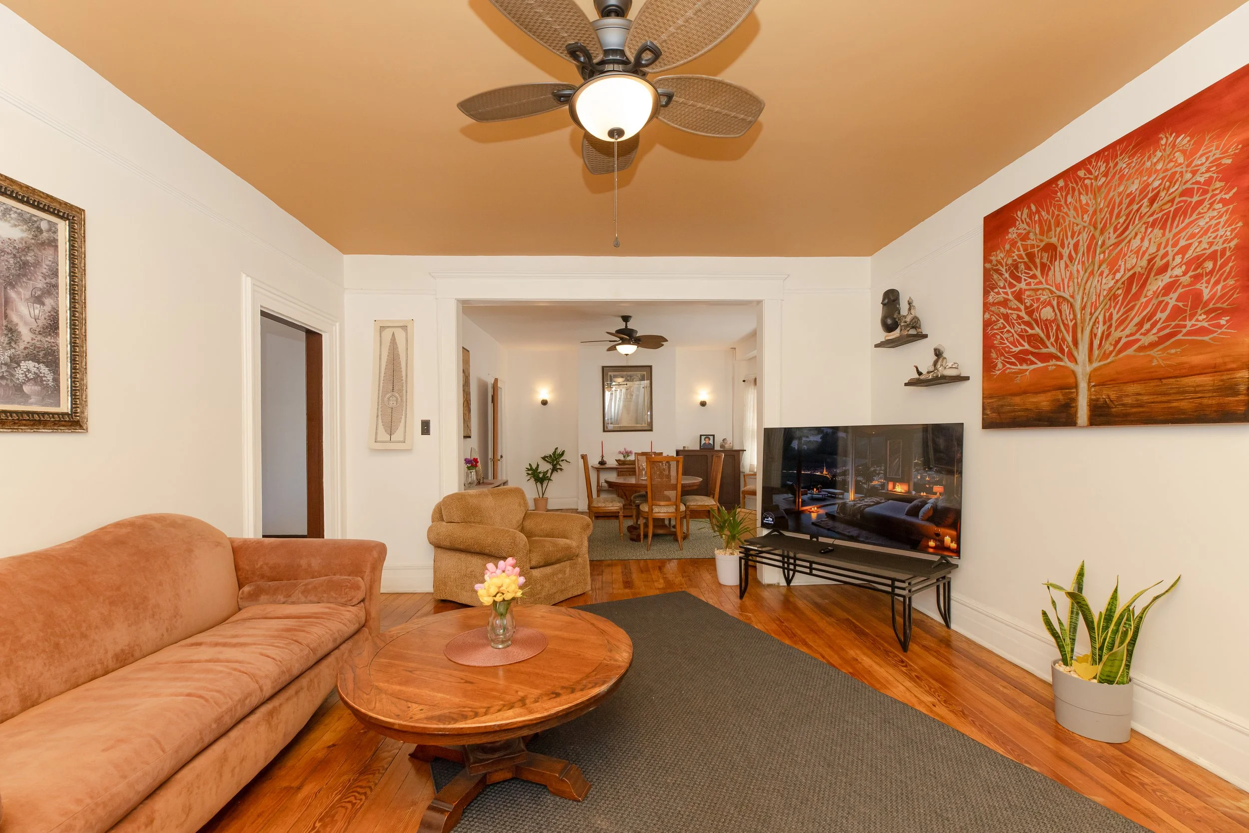 Living room with a peach sofa, a round wooden coffee table with flowers, a beige armchair, a black TV on a black metal stand, wall art, houseplants, and a dining area in the background with a round table and chairs, and a ceiling fan with a light.