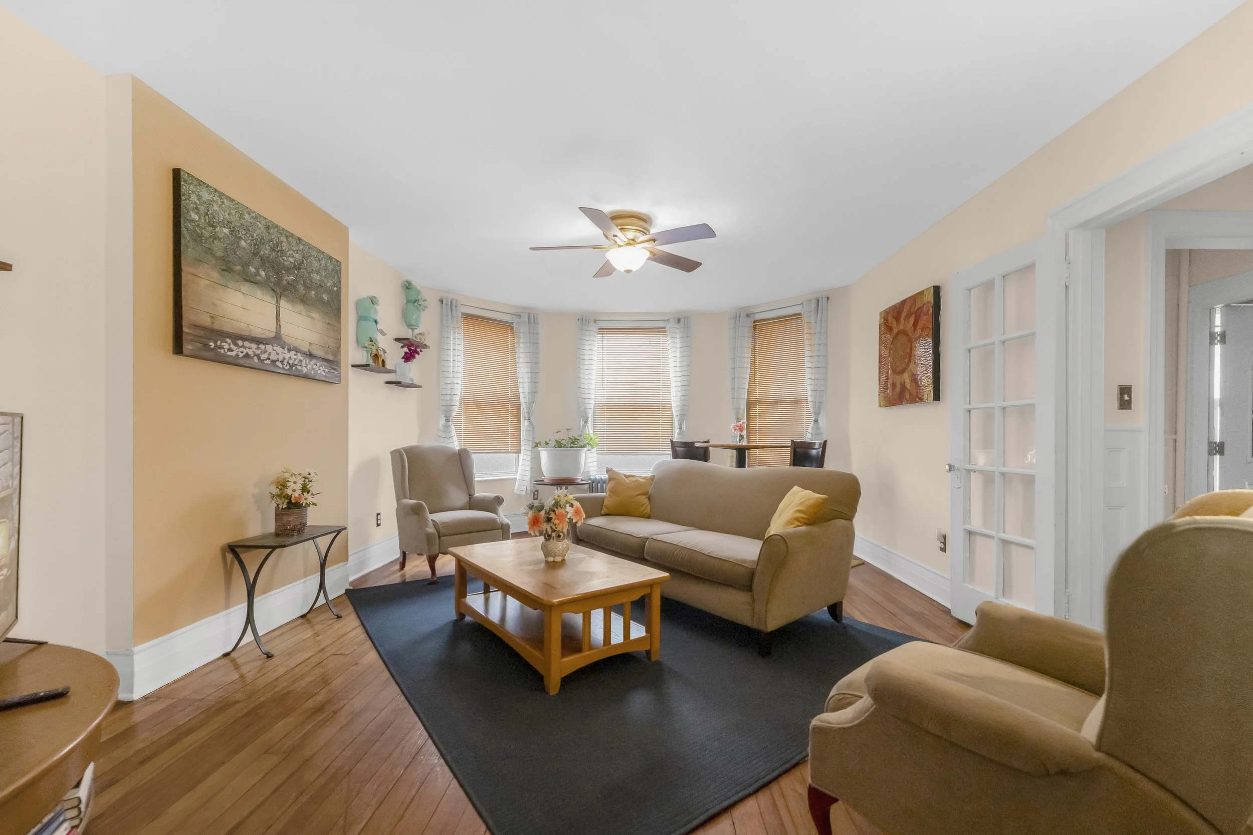 Living room with beige walls, hardwood floors, and a black rug. Contains a beige sofa, an armchair, a coffee table with flowers, and a ceiling fan. Windows with blinds and curtains in the background.