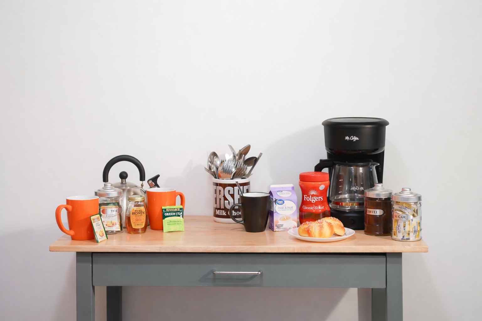 Coffee station with orange mugs, tea, honey, green tea, sugar, coffee maker, cups, and pastries on a wooden table.