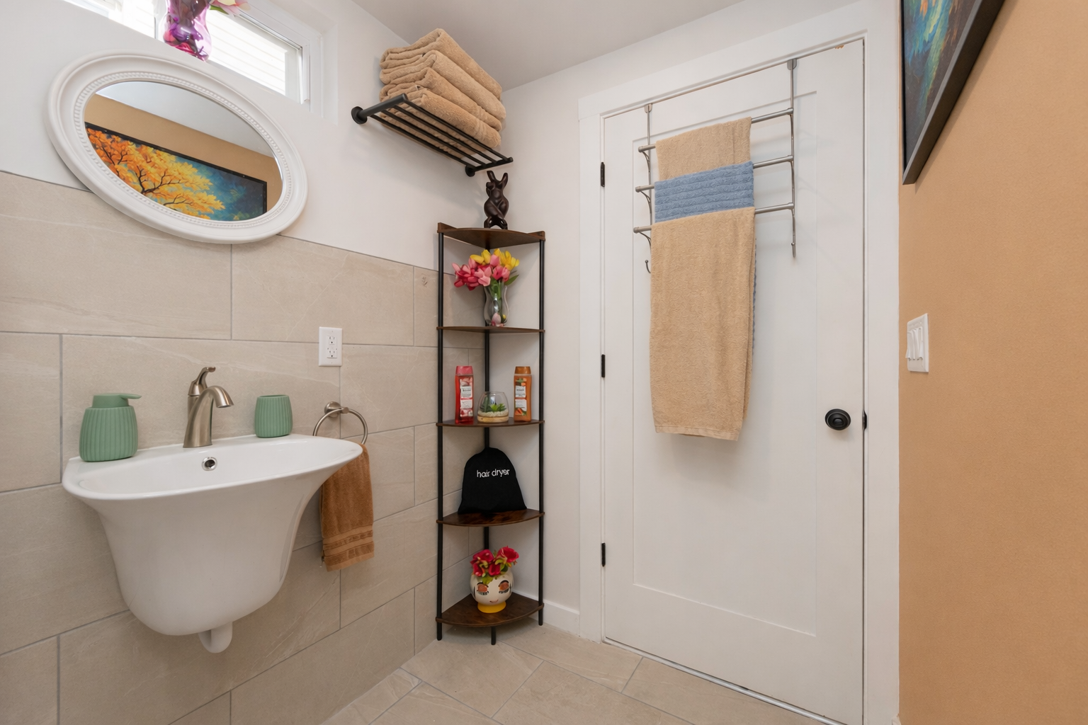 A bathroom with a small wall-mounted sink, a towel hanging from the sink, and a black towel rack above the sink. There are wooden shelves with decorative items, a colorful painting on the wall, and a mirror with a white frame above the sink. The door has a towel rack with a beige and blue towel.