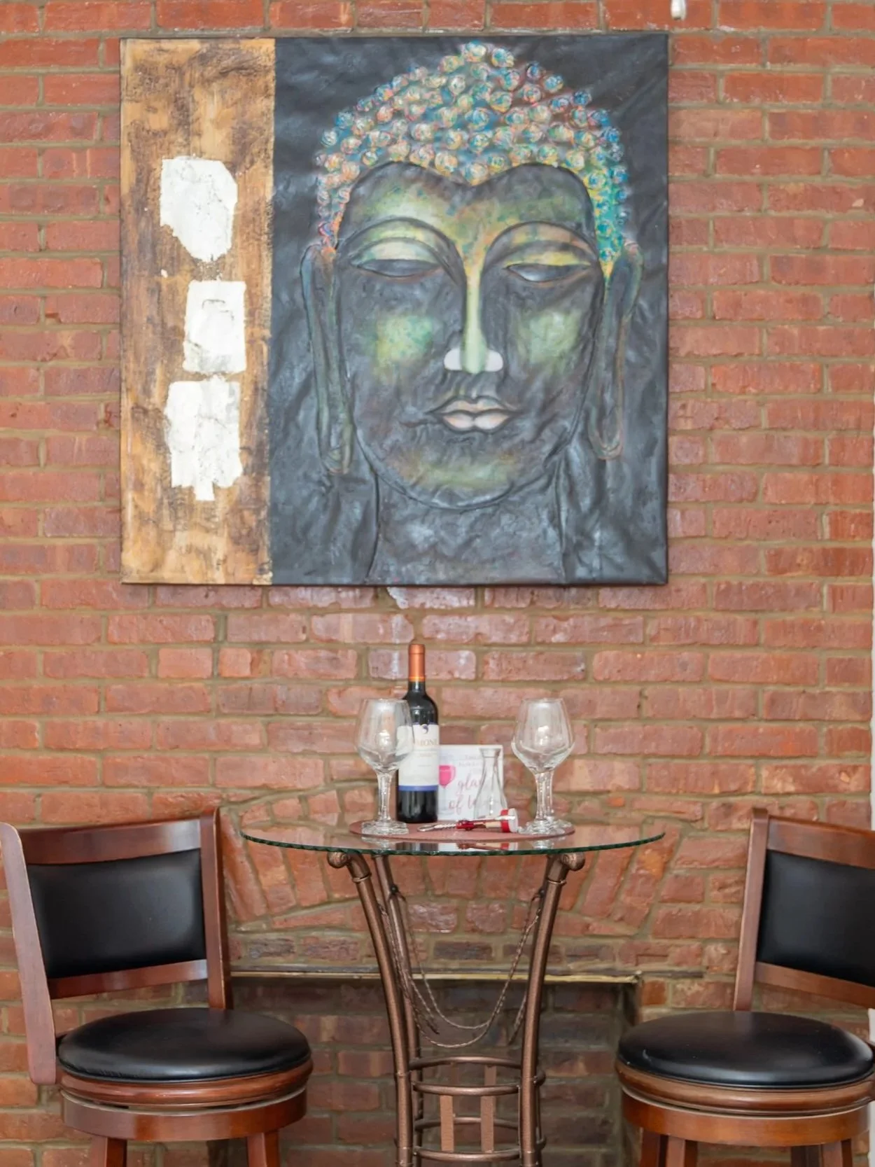 A small round glass table set for two with two empty wine glasses, a bottle of red wine, a wine opener, and a small menu, in front of a brick wall with a large painting of a serene Buddha face.
