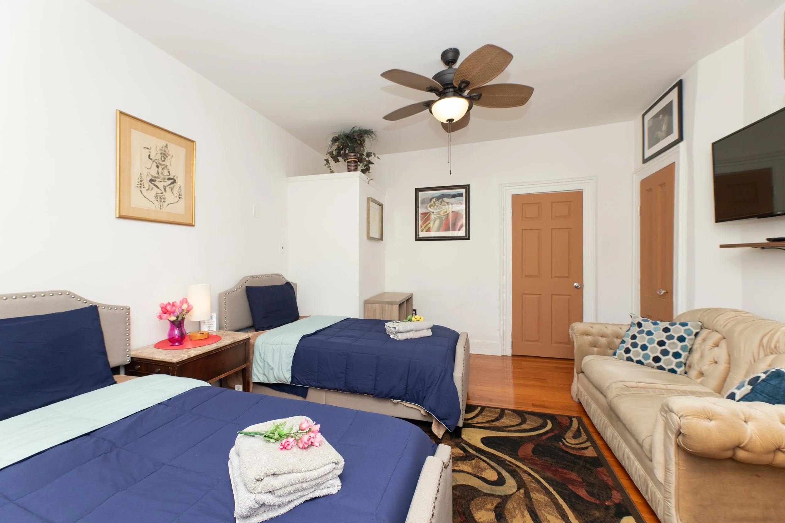Room featuring two beds with navy blue bedding, a beige sofa with patterned pillows, a wall-mounted TV, wooden door, ceiling fan, and framed artwork.