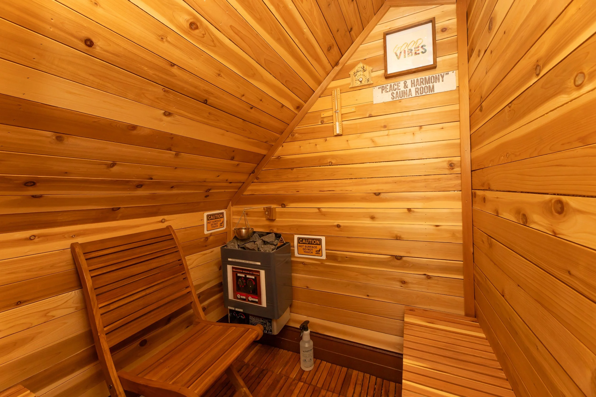 Wood-paneled sauna room with a small heater filled with stones, two caution signs, and a wooden chair. There are framed signs on the wall, one says 'Good Vibes' and another indicates 'Peace & Harmony Sauna Room'. A spray bottle is on the floor.