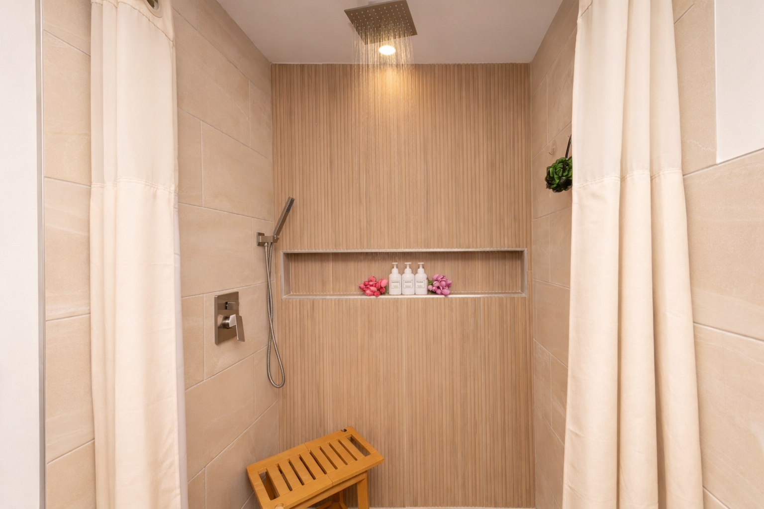 A modern shower with beige tiles, a rainfall showerhead, a handheld shower, a built-in shelf with soap and pink flowers, and a wooden stool, enclosed by cream-colored curtains.