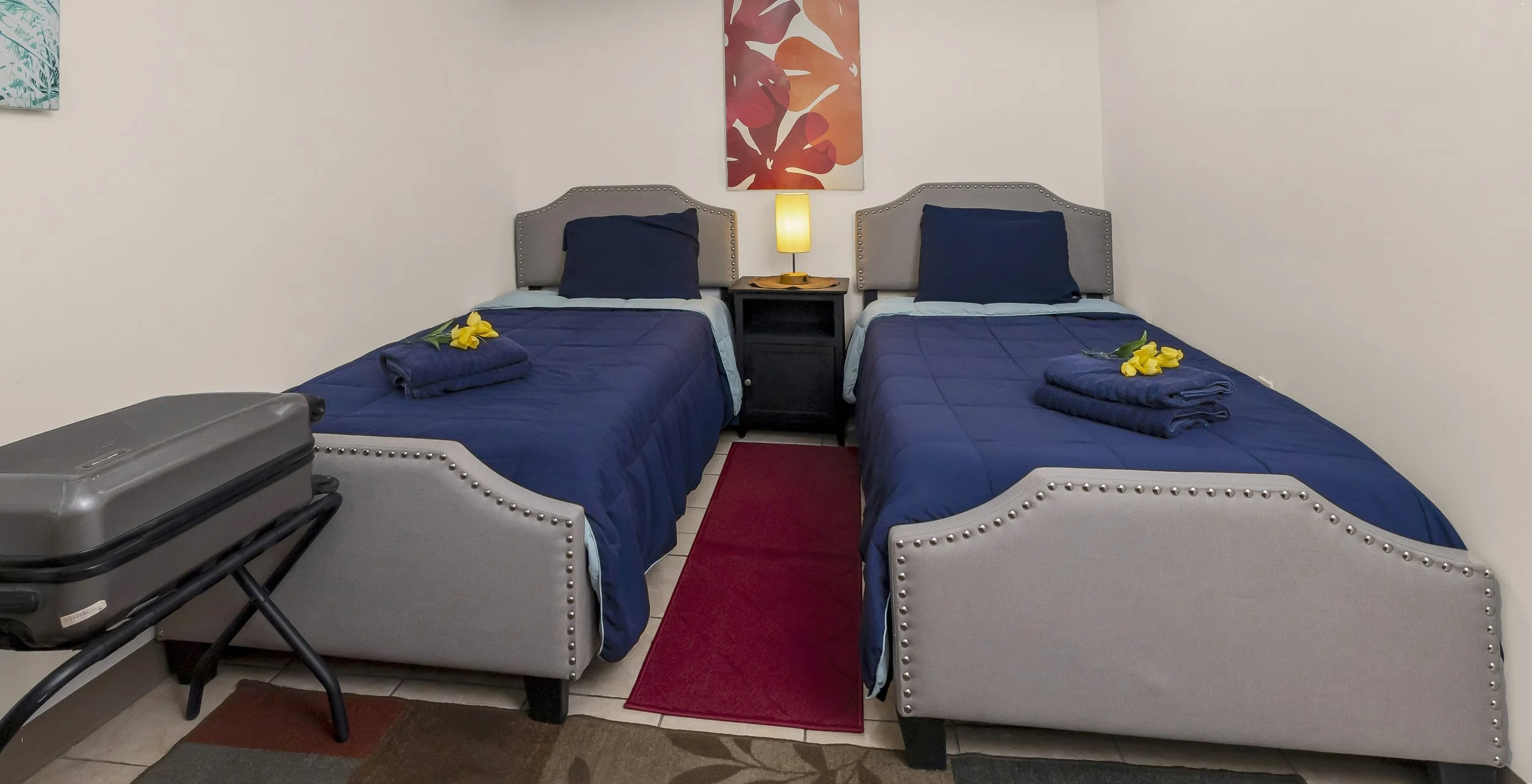 A hotel room with two single beds covered in dark blue bedding, each with a rolled towel and yellow flowers on top. There is a nightstand between the beds with a lamp on it, and a piece of artwork above the beds. An additional suitcase is on a luggage rack near the foot of one bed. A red rug runs between the beds on the tiled floor.