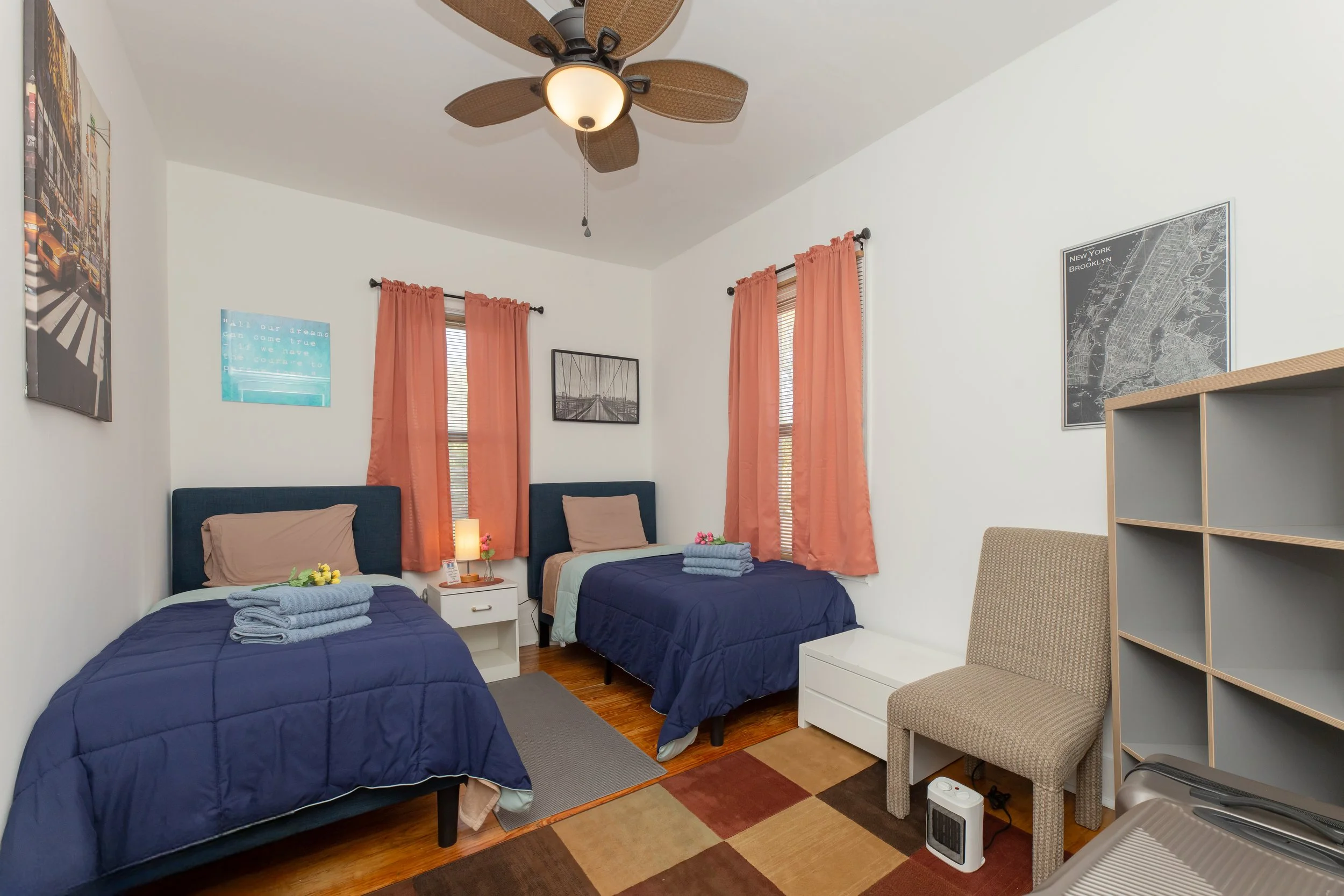 Bedroom with two twin beds with blue bedding, beige pillows, and folded towels on each bed, nightstand with lamp, two windows with pink curtains, wall art, a beige chair, and an overhead ceiling fan.