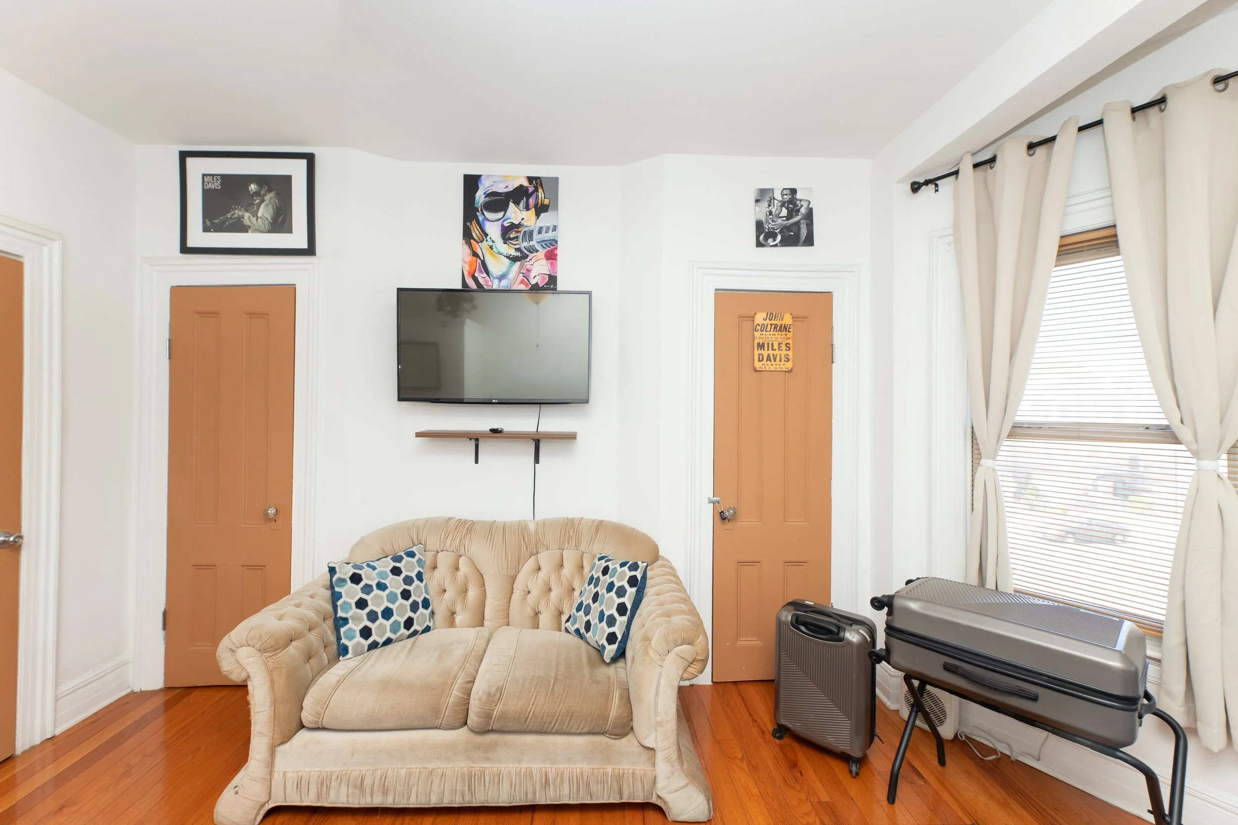 Living room with beige tufted couch, two blue and white patterned pillows, hardwood floor, two brown doors, a wall-mounted TV, framed photos and artwork above, a window with cream curtains, and a small luggage set.