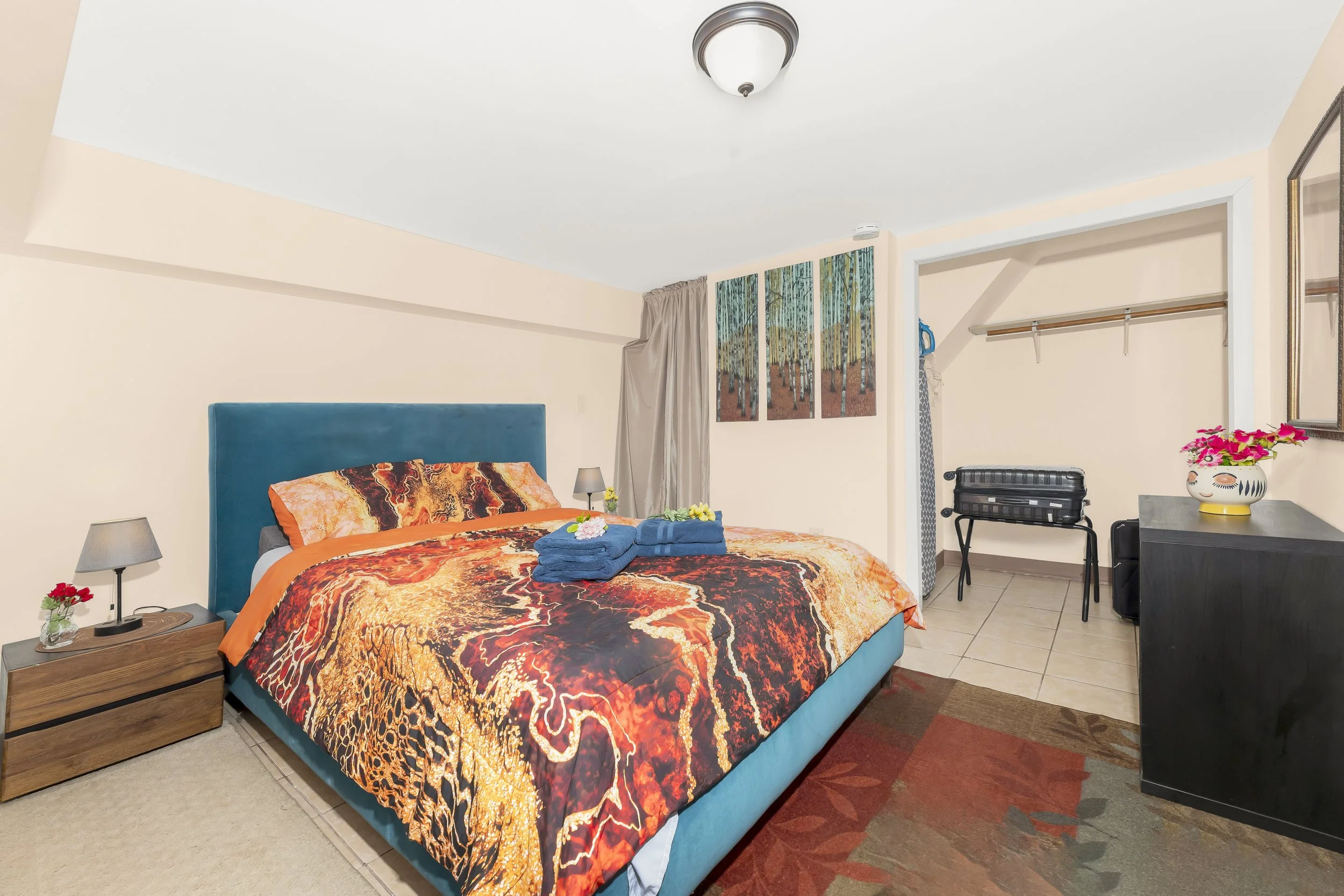Hotel room featuring a blue bed with colorful animal print bedding, two bedside lamps, a small table with a flower vase, and a dresser with pink flowers. The room has a closet with hanging space, a folding luggage rack, a mirror, and wall art depicting a forest scene.