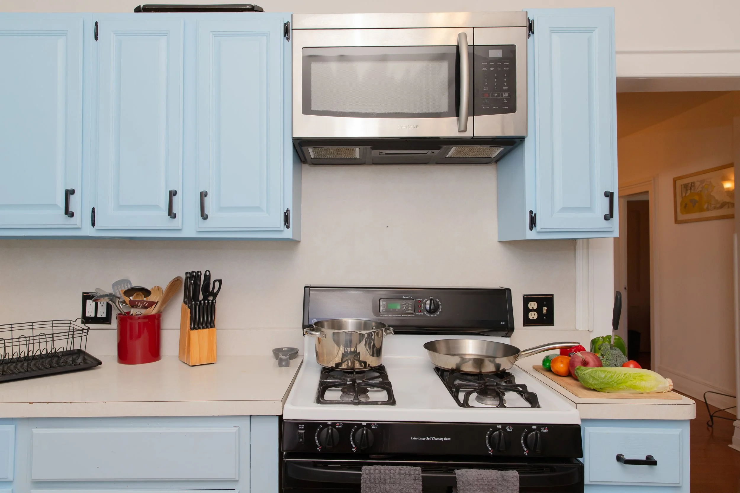 Kitchen with blue cabinets, a stainless steel microwave above the stove, a gas range with two pans, a cutting board with vegetables, and kitchen utensils in containers.