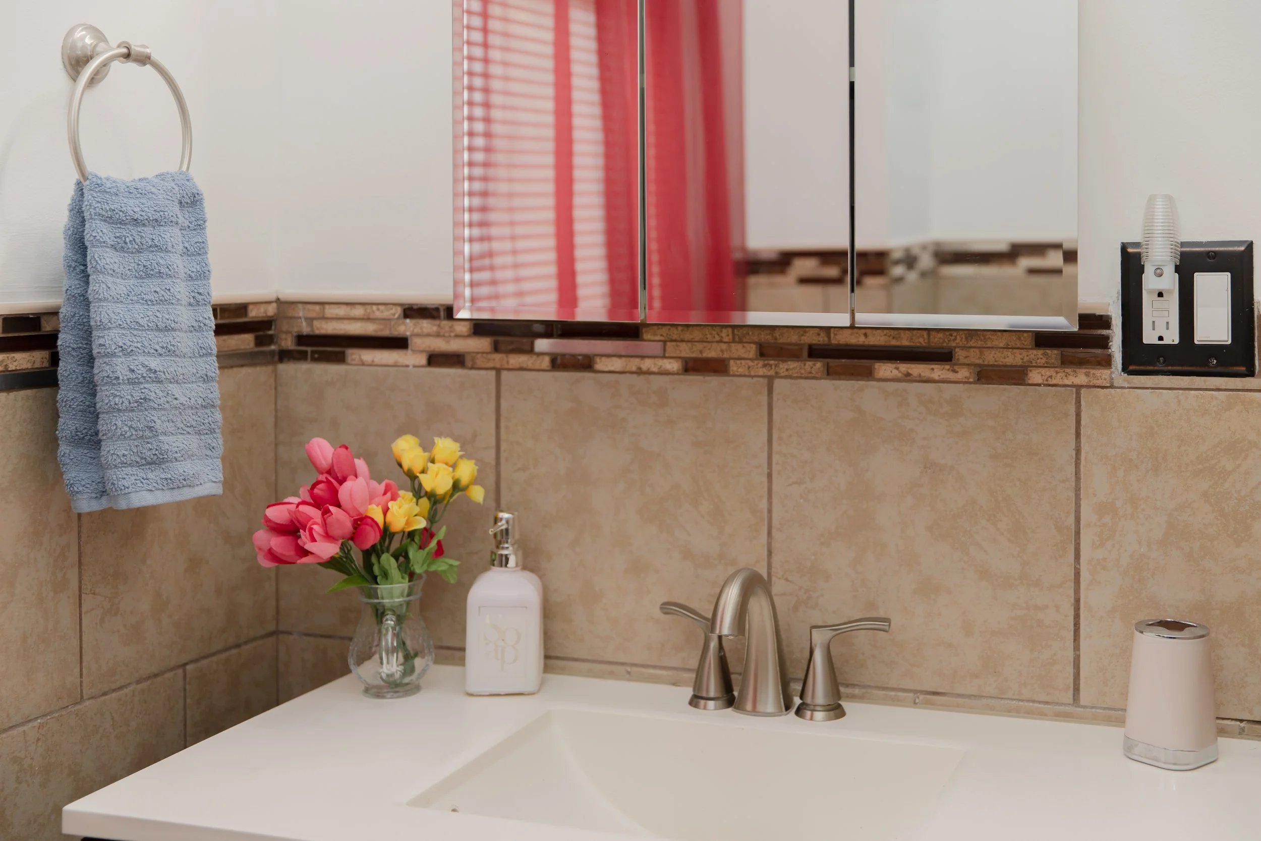 Bathroom sink with a vase of pink and yellow artificial flowers, a soap dispenser, a toothbrush holder, a blue towel on a ring, and a mirror above the sink.
