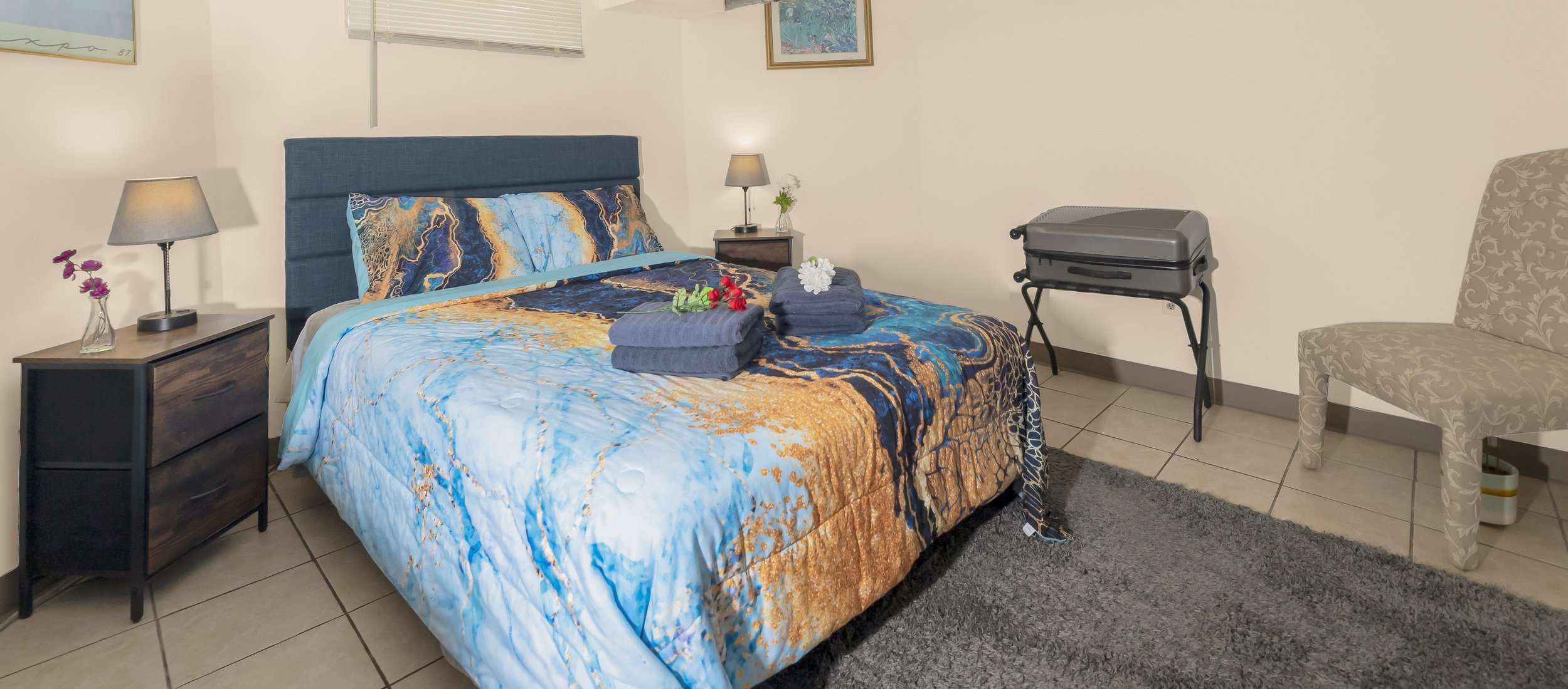 Hotel room with a bed that has a blue and gold patterned bedspread, two rolled towels with flowers on top, two bedside tables with lamps, artwork on the wall, a beige patterned chair, a small grill, and a gray rug on a tiled floor.