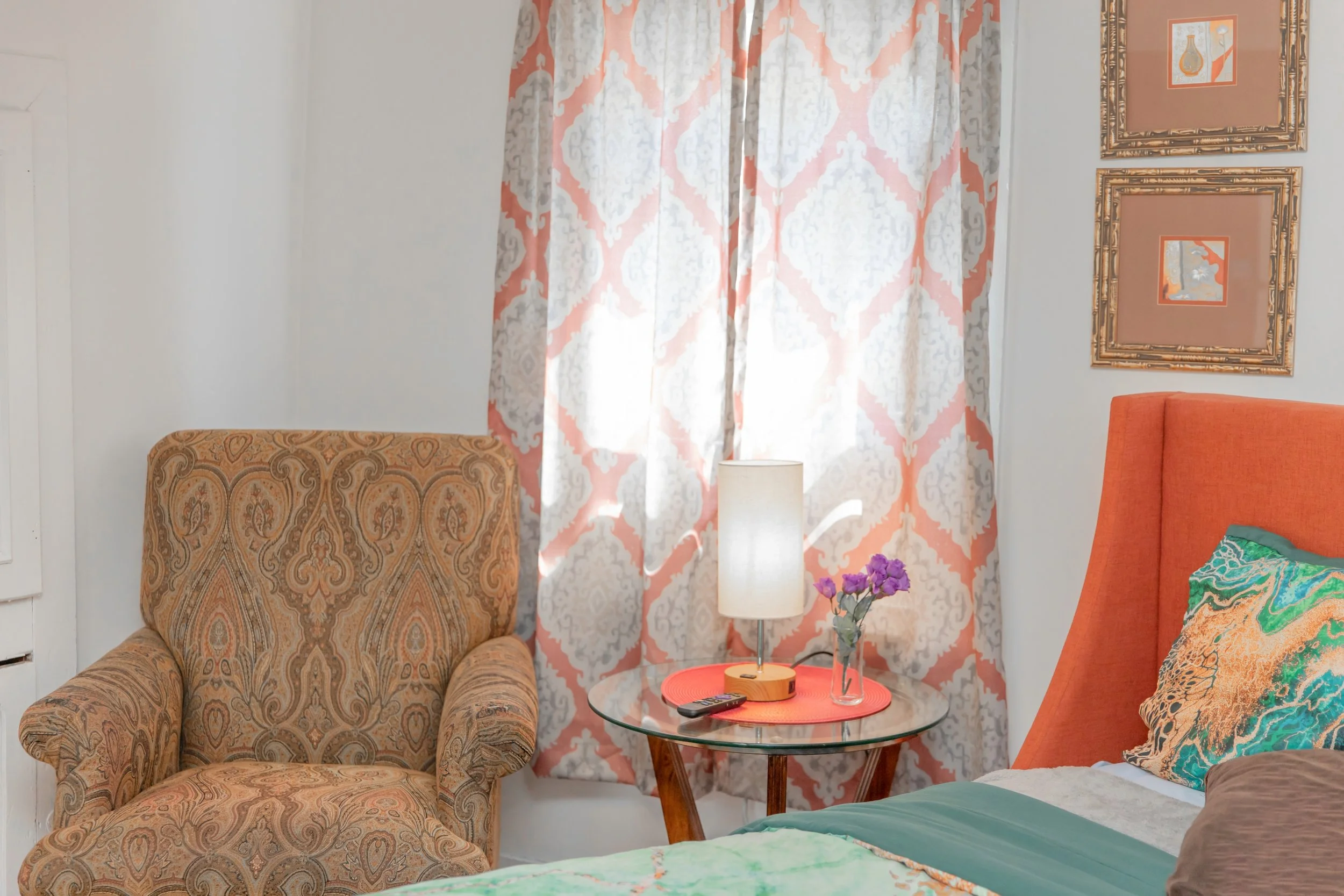 A cozy bedroom corner with an upholstered armchair, a glass-topped nightstand, a table lamp, a flower vase with purple flowers, and patterned curtains with pink and white designs. Part of a bed with an orange headboard and decorative pillows is visible.