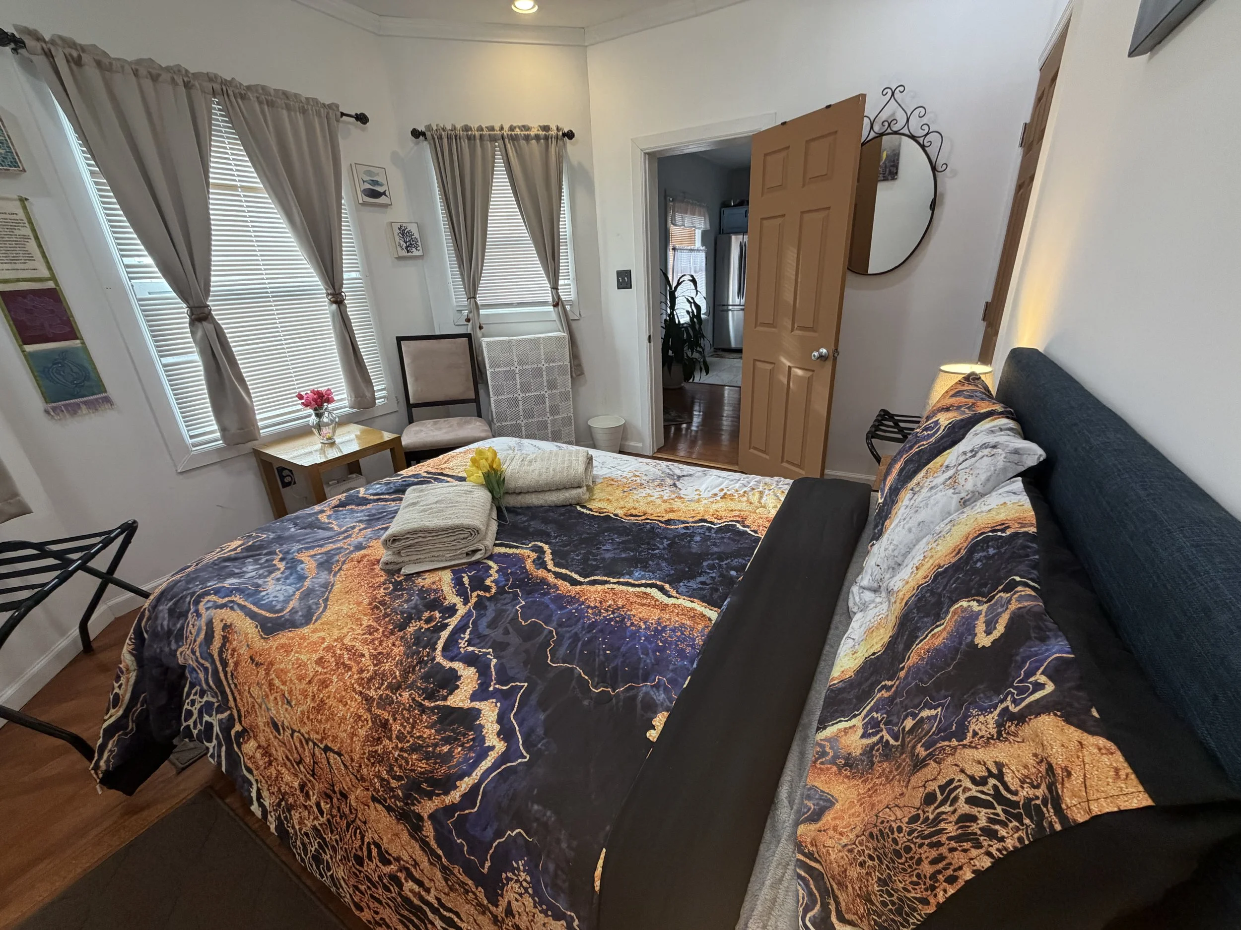 A bedroom with a bed covered by a colorful blanket featuring orange, yellow, and black abstract patterns. Towels and a flower are placed on the bed. The room has three windows with beige curtains, some framed artwork on the walls, a chair, and a small table with a pink flower. An open door reveals a view into a kitchen with a refrigerator and a plant.