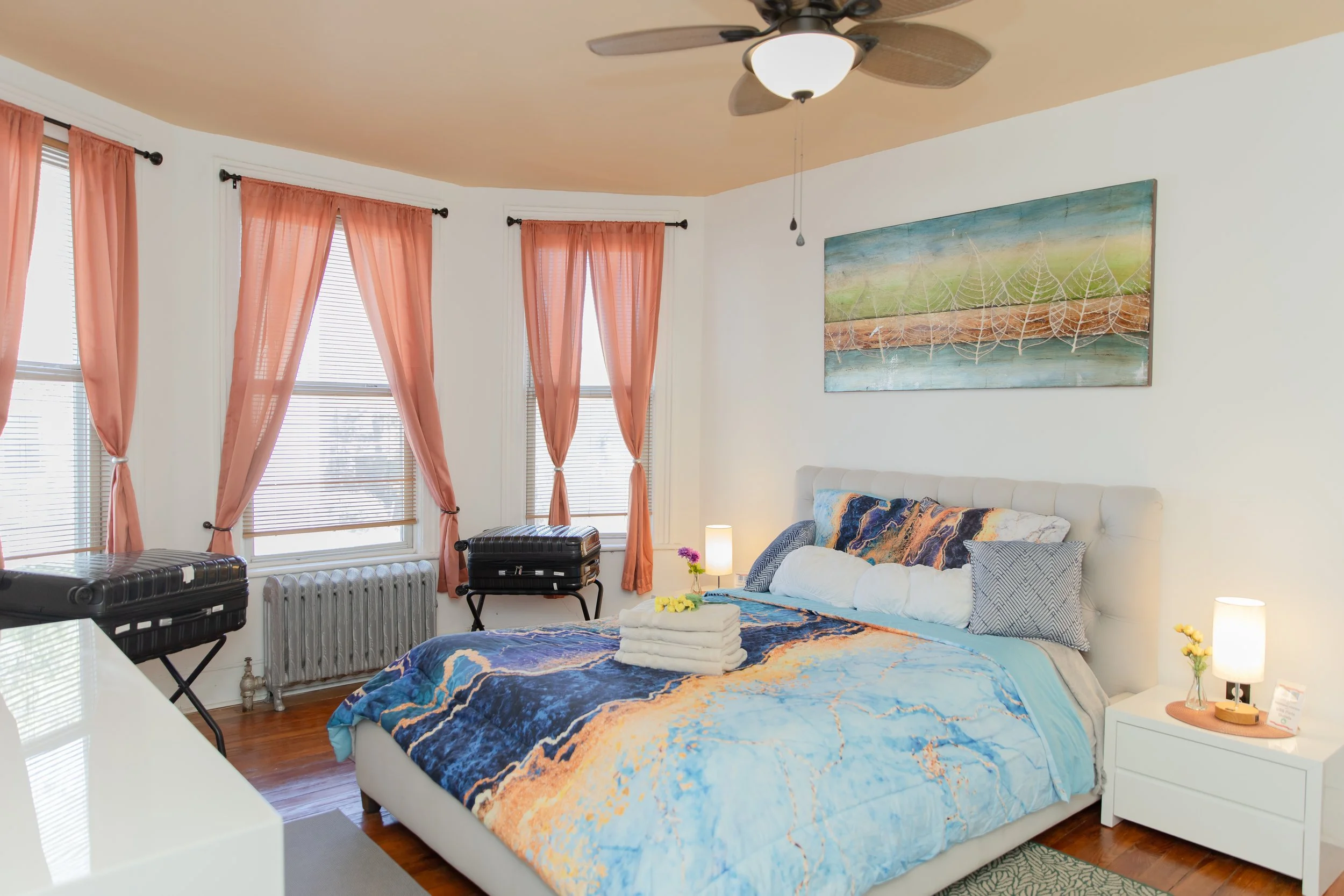 A bedroom with a white bed, colorful bedspread, and pillows. Three windows with pink curtains, a ceiling fan, and artwork of spider webs on the wall. Nightstand with a lamp, flowers, and a note. Suitcases on stands by the windows.