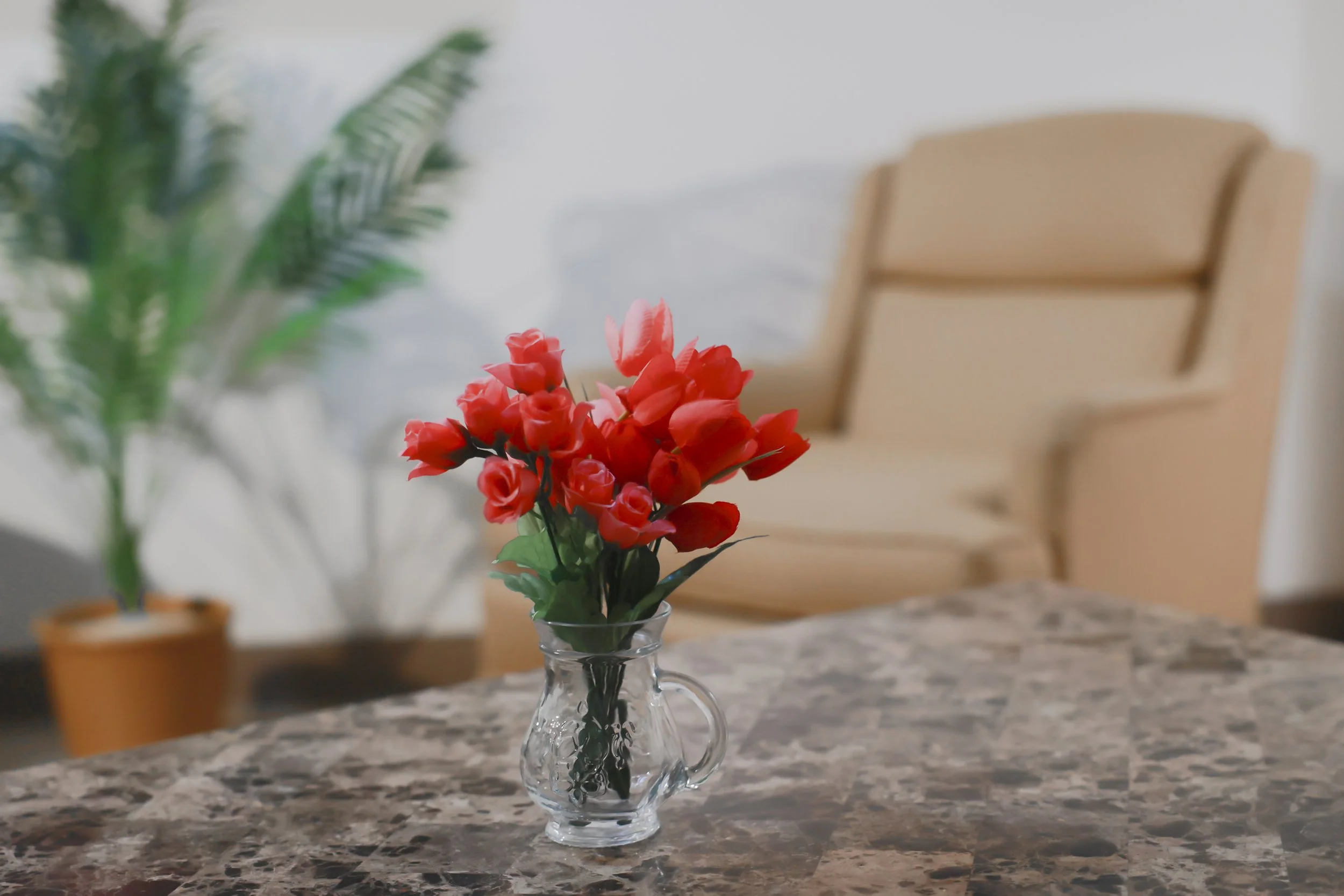 Yellow chair, green potted plant, and pink flowers in a glass vase on a marble table in a room.