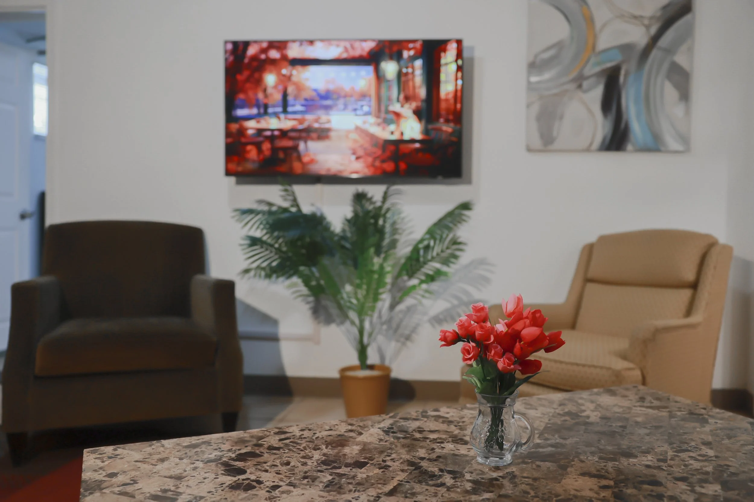 Living room with two armchairs, a pot with green plant, a wall-mounted TV displaying an autumn scene, abstract wall art, and a table with a glass vase of pink flowers.