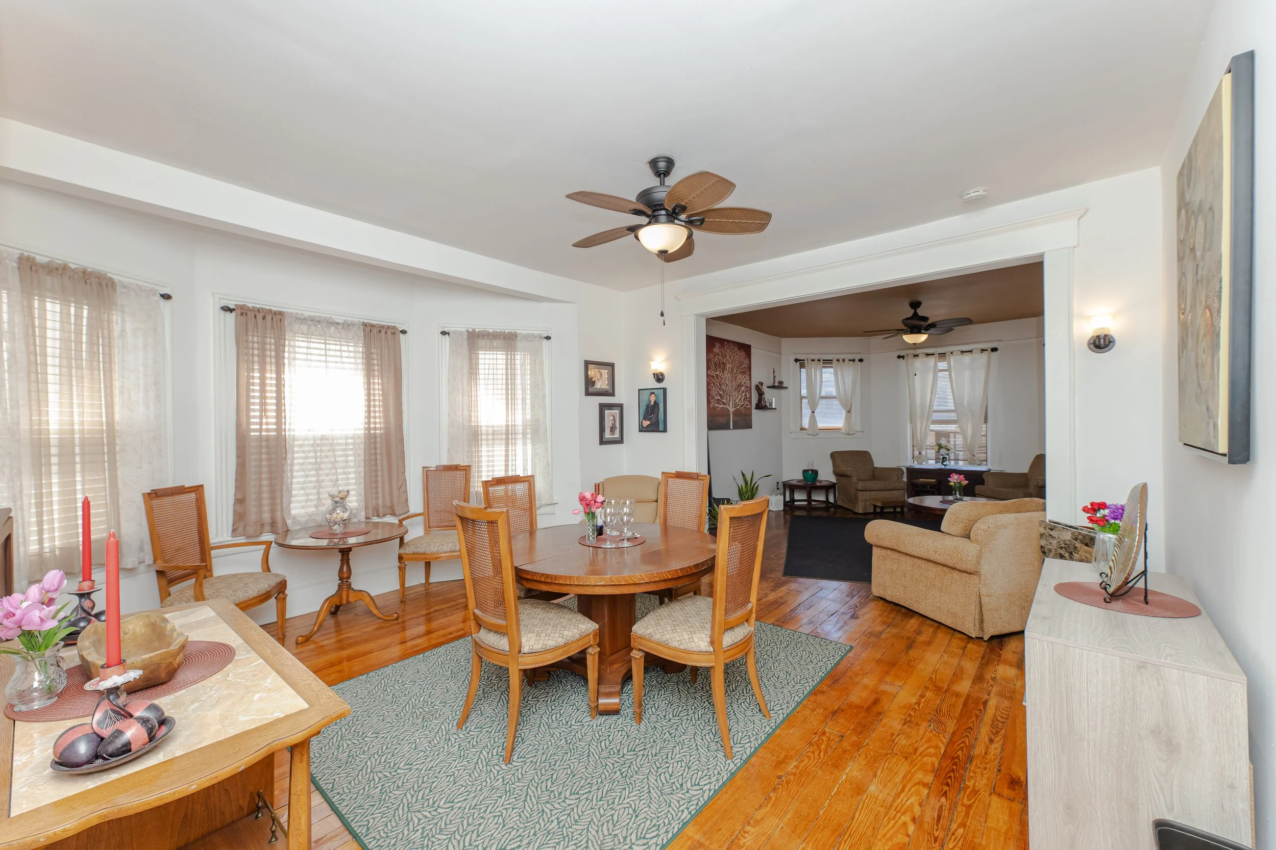 Living and dining room with hardwood floors, beige and tan furniture, and multiple windows with curtains. Dining area features a round wooden table with six chairs, and a sideboard with pink candles and flowers. The living room has sofas, armchairs, and artwork on the walls.