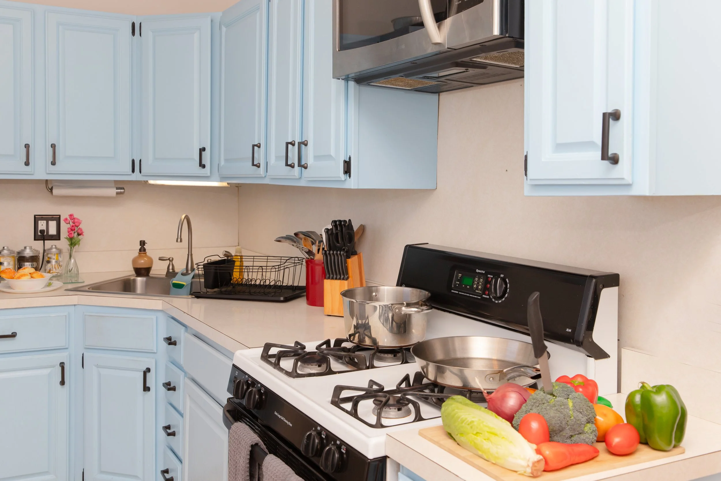 Kitchen with blue cabinets, a stove with a pot, a pan, and fresh vegetables on a cutting board, including a green bell pepper, broccoli, tomatoes, a red onion, and carrots.