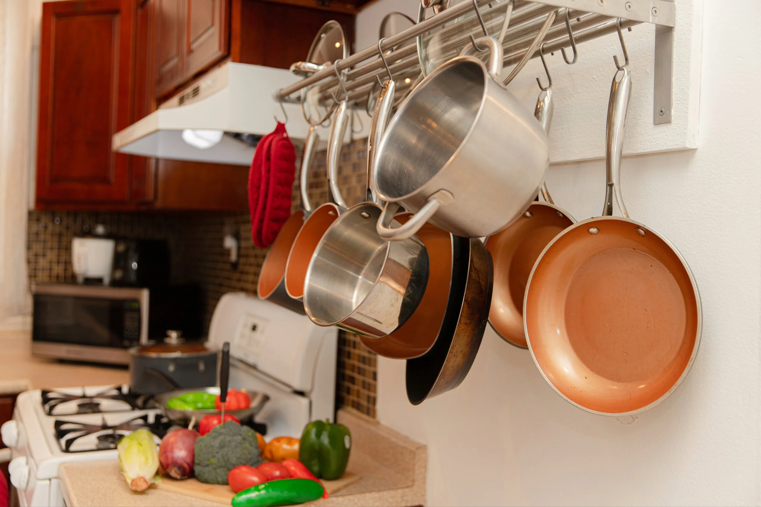 Hanging pots and pans above a kitchen countertop with fresh vegetables like broccoli, tomatoes, and bell peppers.