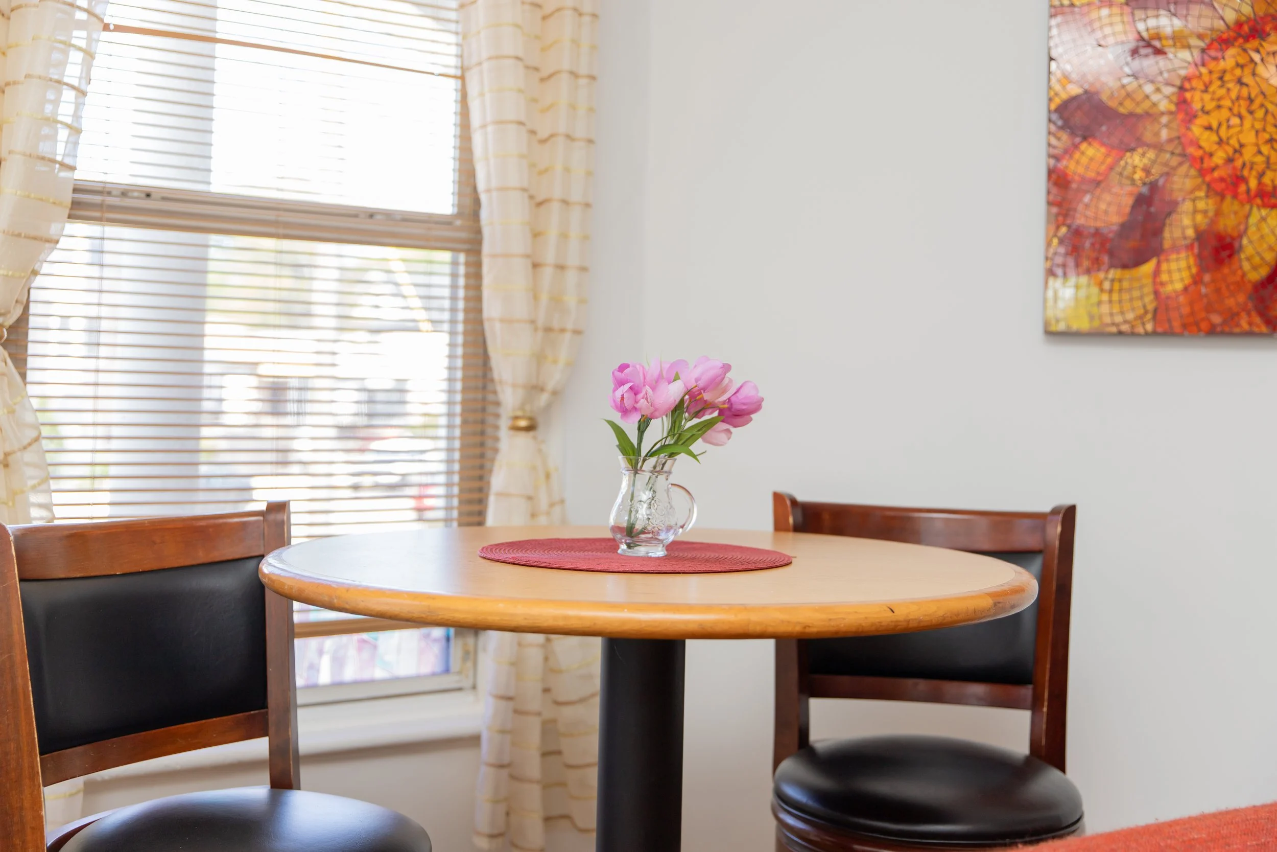 A round wooden table with a red placemat and a glass vase with pink flowers, placed near a window with light-colored curtains and blinds, in a room with white walls and colorful artwork.