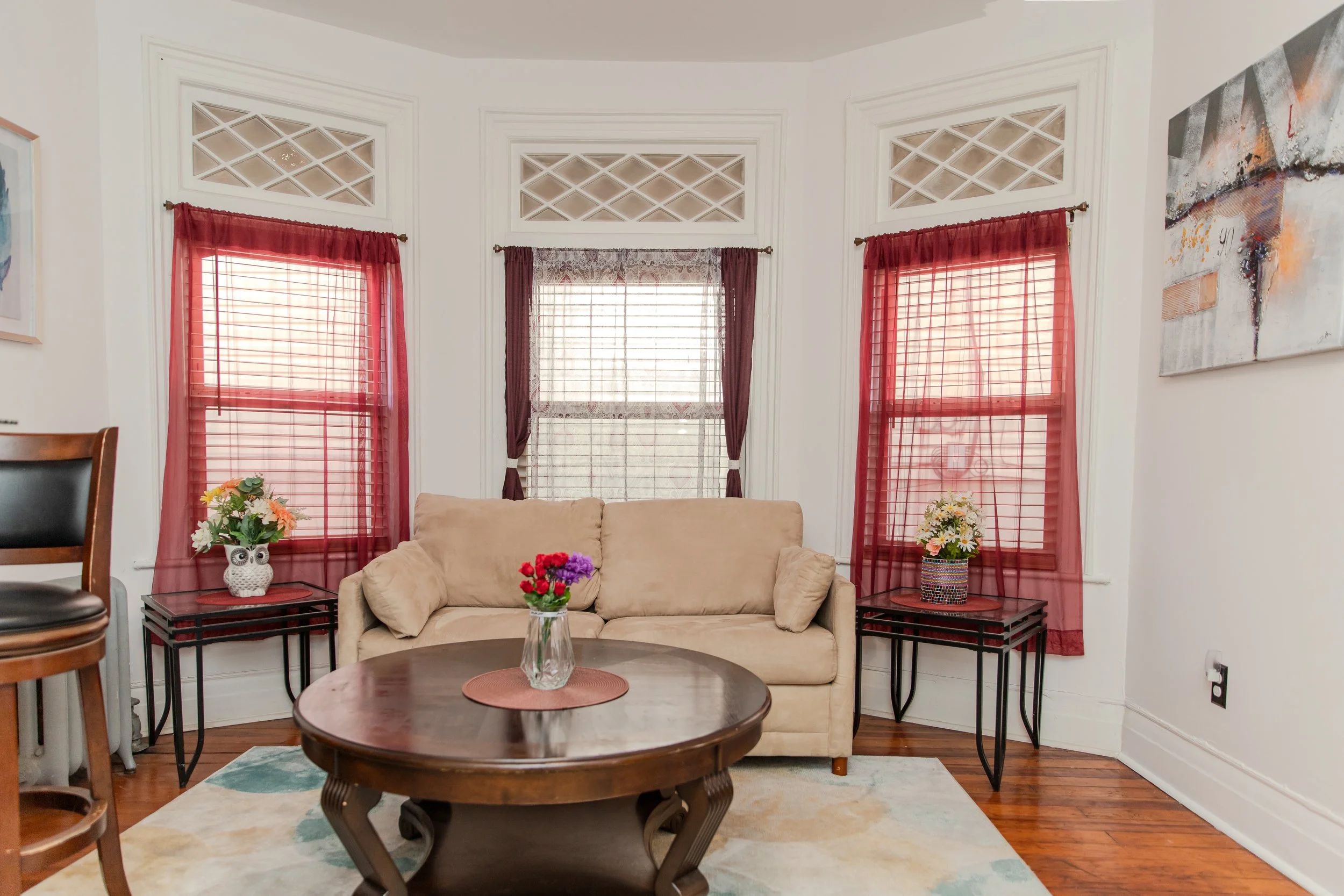 Living room with three windows with red and patterned curtains, beige sofa, wooden coffee table with a flower vase, and two black side tables with flower arrangements.