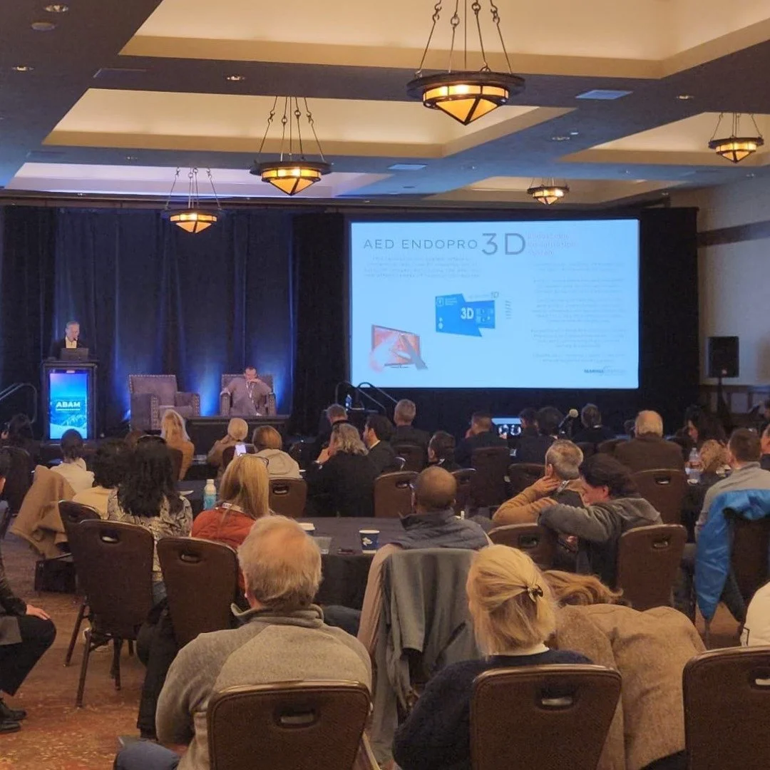 A conference room with audience members seated at round tables, a stage with a speaker at a podium, and a large presentation screen displaying a slide about AED Endopro 3D.