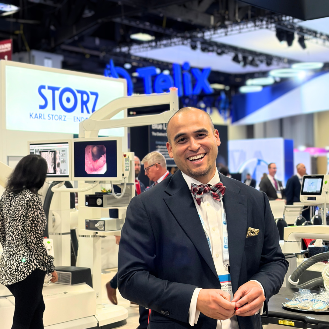 A smiling man in a suit and bow tie at a medical technology conference, surrounded by medical equipment and display screens.