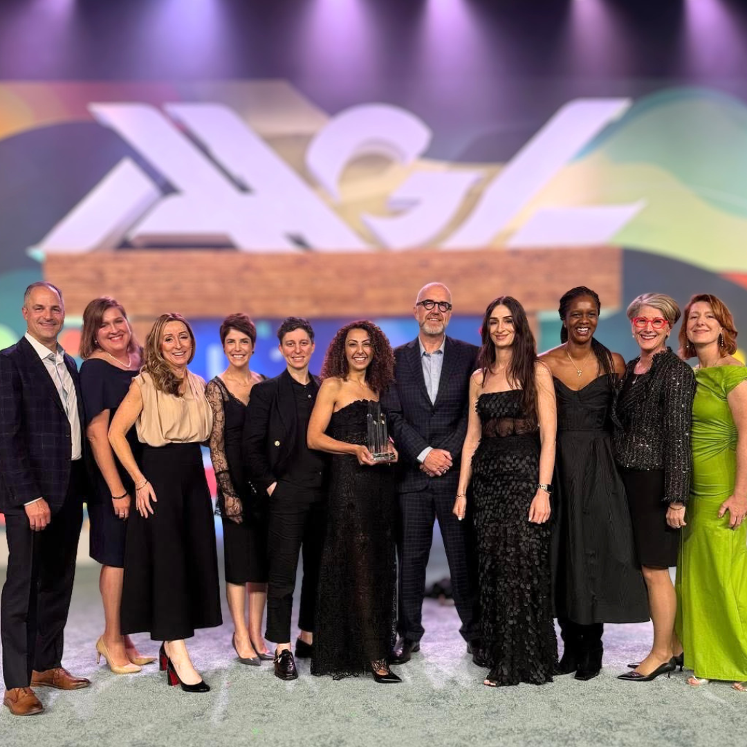 Group of eleven people dressed in formal attire standing on stage, holding an award, with colorful background and large logo projected behind them.
