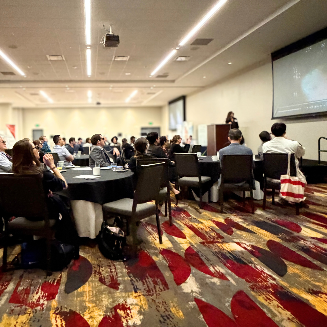 A conference room filled with people seated at round tables, listening to a woman speaking at a podium near a large screen.