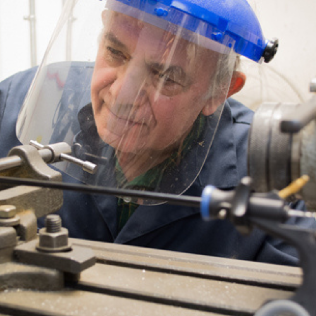 A man working with machinery in a workshop, wearing a blue safety helmet and a transparent face shield.