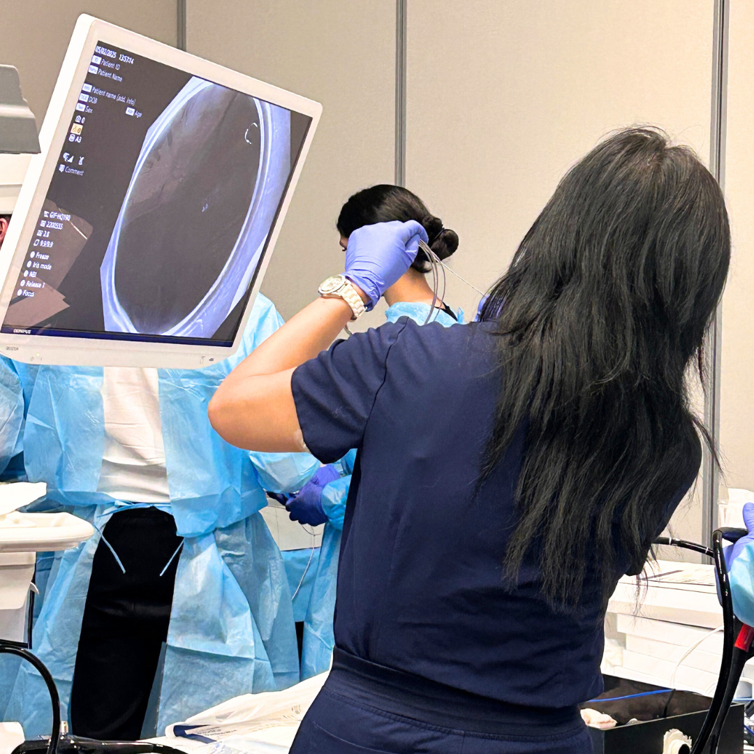 Healthcare workers in scrubs and masks perform a medical procedure in front of an ultrasound screen. One worker holds a medical tool near a patient, who is partly visible. The ultrasound monitor displays an internal scan.