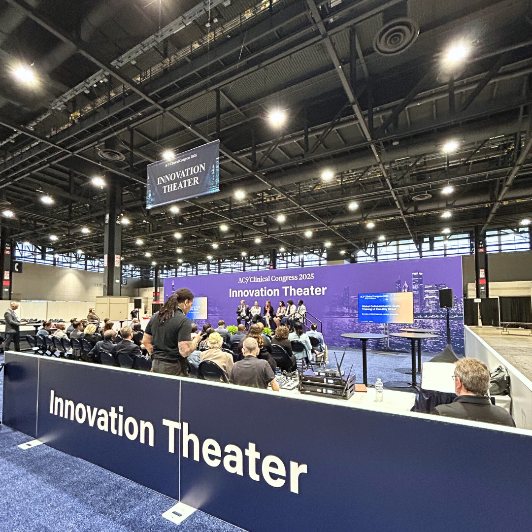 A conference or event in a large indoor space with an elevated stage labeled 'Innovation Theater.' Several panelists are sitting on the stage, and an audience is seated facing them. The background has a purple banner with white text showing 'ACS/Clinical Congress 2025' and 'Innovation Theater,' with cityscape graphics. There is a screen on the right side displaying a presentation, and some equipment and water bottles are visible.