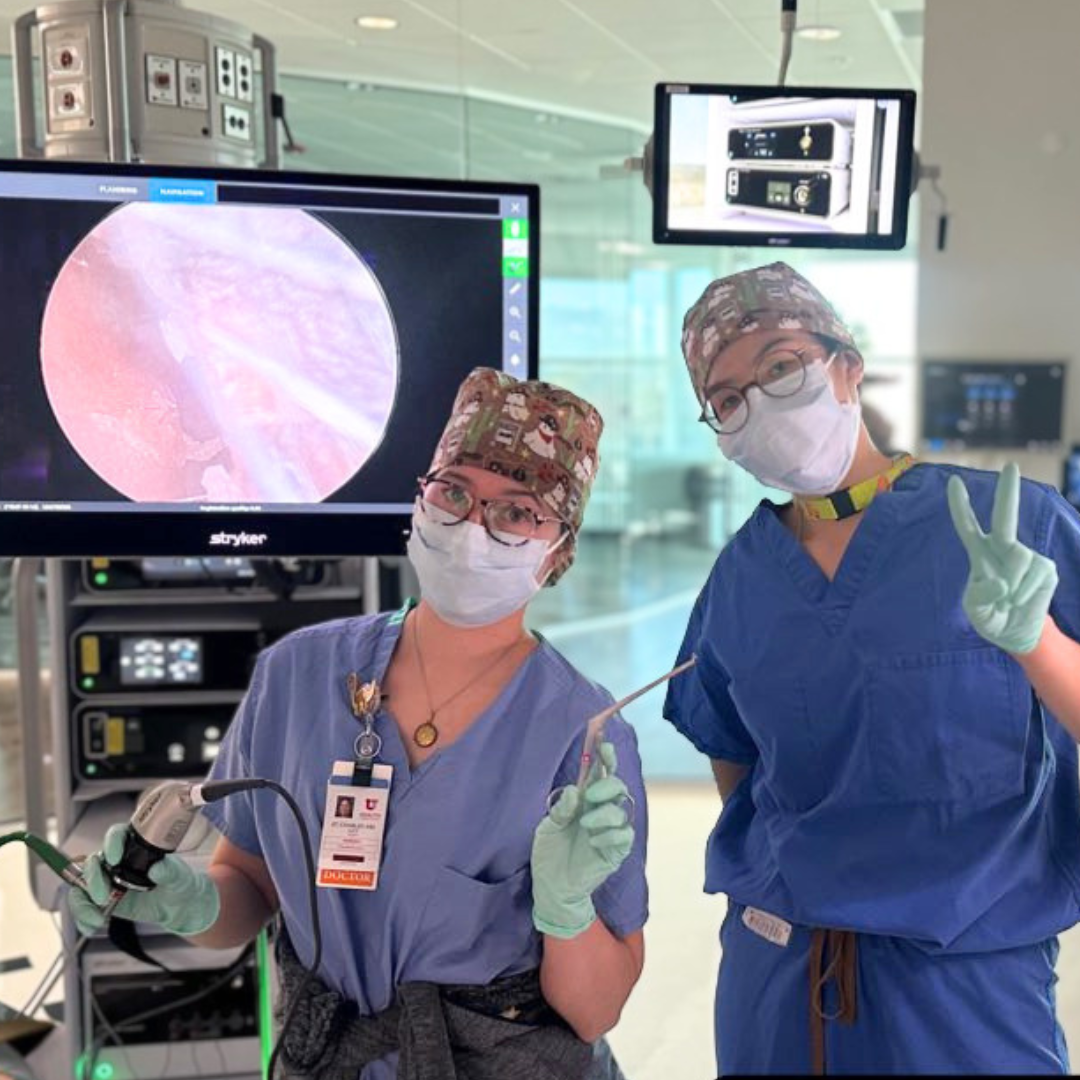 Two medical professionals in blue scrubs and face masks in a hospital operating room, with one making a peace sign and the other holding a medical instrument, behind a large monitor displaying an endoscopic view.