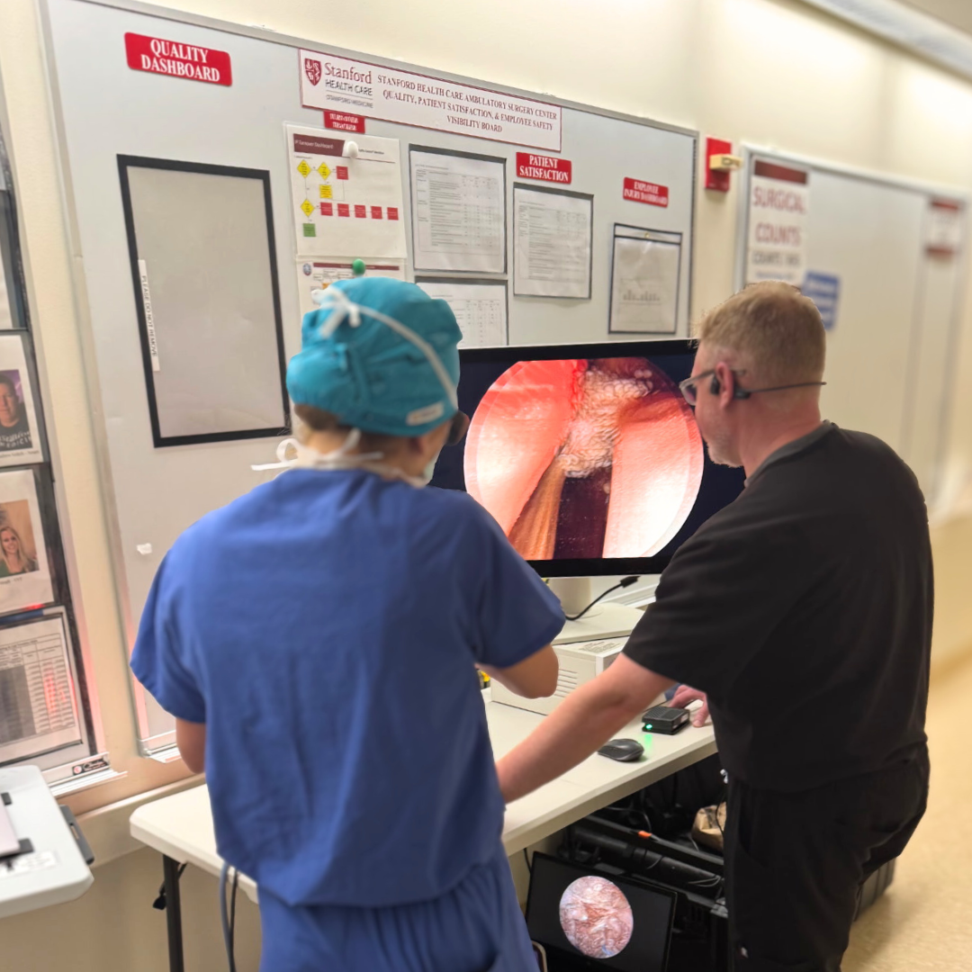 Two medical professionals, one in blue scrubs and surgical cap and the other in black scrubs and glasses, examining a medical image on a monitor in a hospital setting. The monitor displays an endoscopic view of a patient’s internal anatomy.