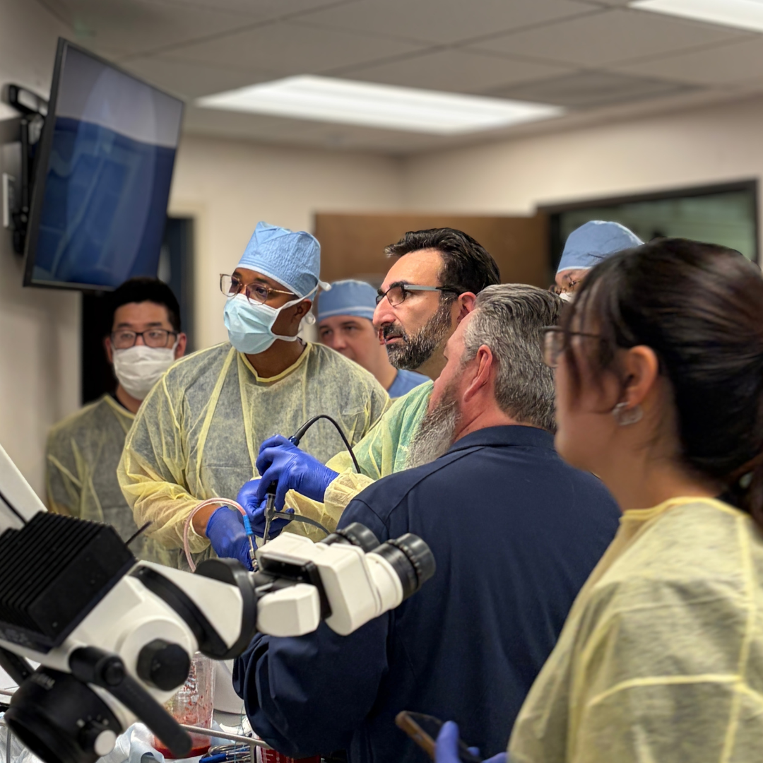 Medical professionals, including surgeons and students, are gathered around a microscope and a patient, participating in a surgical procedure or medical training session in a hospital or clinic setting.