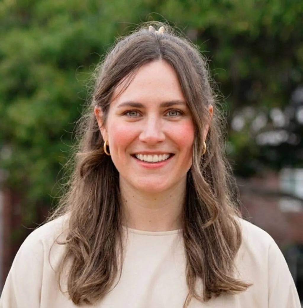 A woman with long brown wavy hair, fair skin, and blue eyes, smiling outdoors with greenery in the background, wearing a beige top and gold hoop earrings.