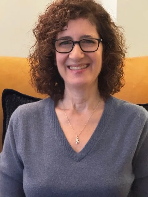 A woman with curly brown hair, wearing glasses, a grey sweater, and a necklace, smiling while sitting on a yellow couch.