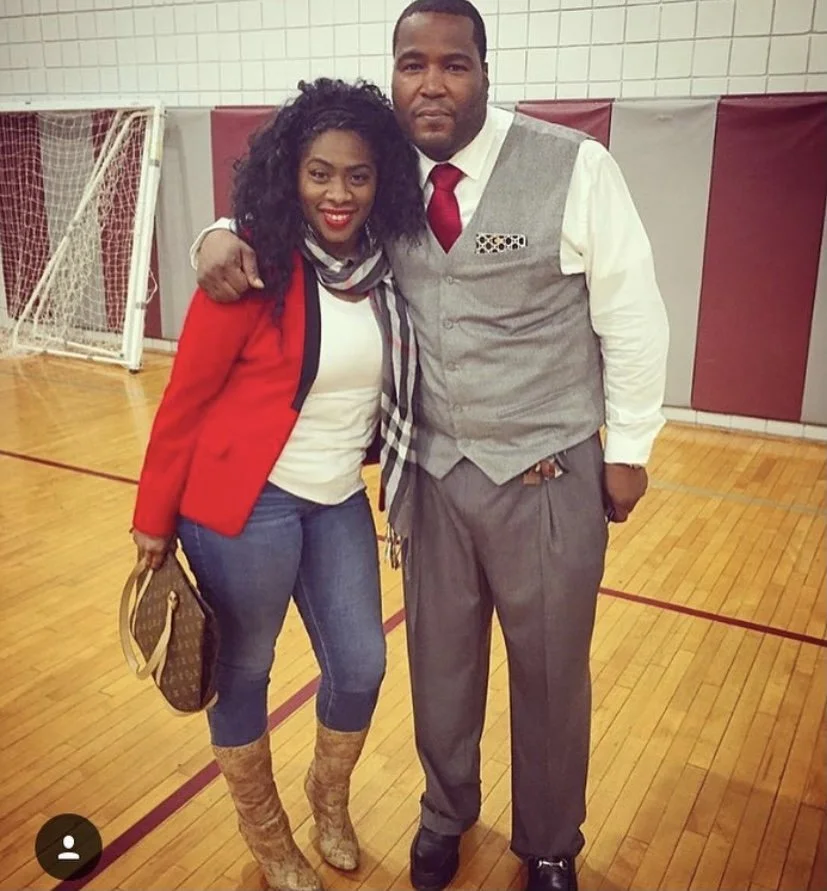 A woman and man standing in a gymnasium, posing for a photo. The woman has curly black hair, red lipstick, a red blazer, white top, blue jeans, and brown patterned boots. The man has short black hair, a white shirt, gray vest, gray trousers, and a re