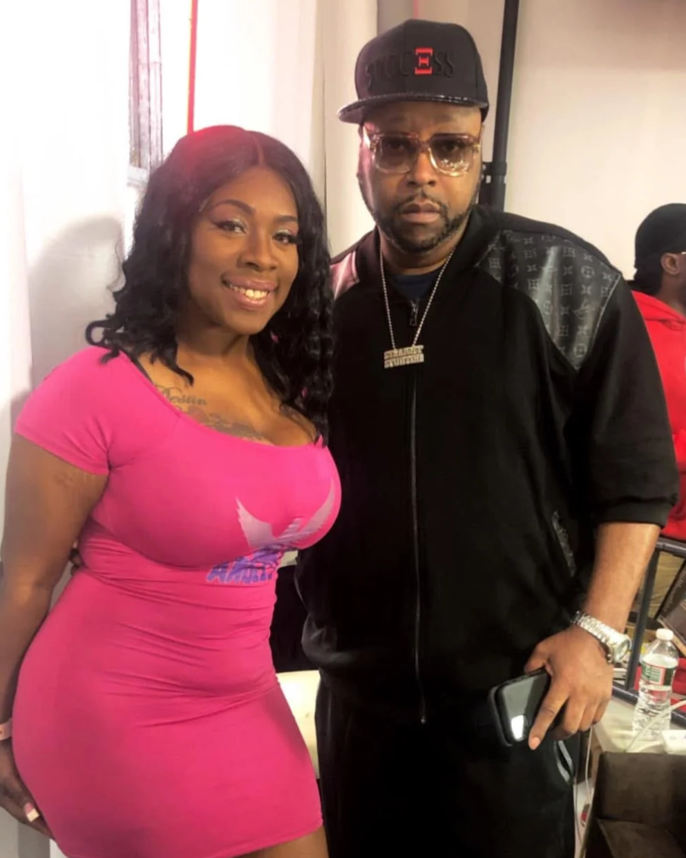 A smiling woman in a bright pink dress stands next to a serious man in black clothing, wearing a cap and glasses, in an indoor setting.