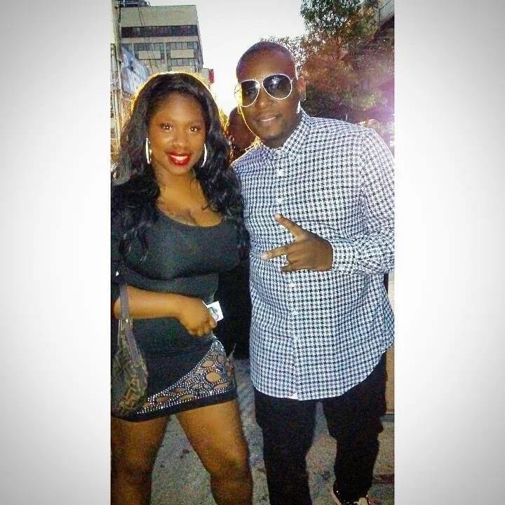 A man and woman posing together outdoors in an urban setting during the evening, with buildings and trees visible in the background. The woman is wearing a black dress with cut-out details, large hoop earrings, and has wavy black hair. The man is wea