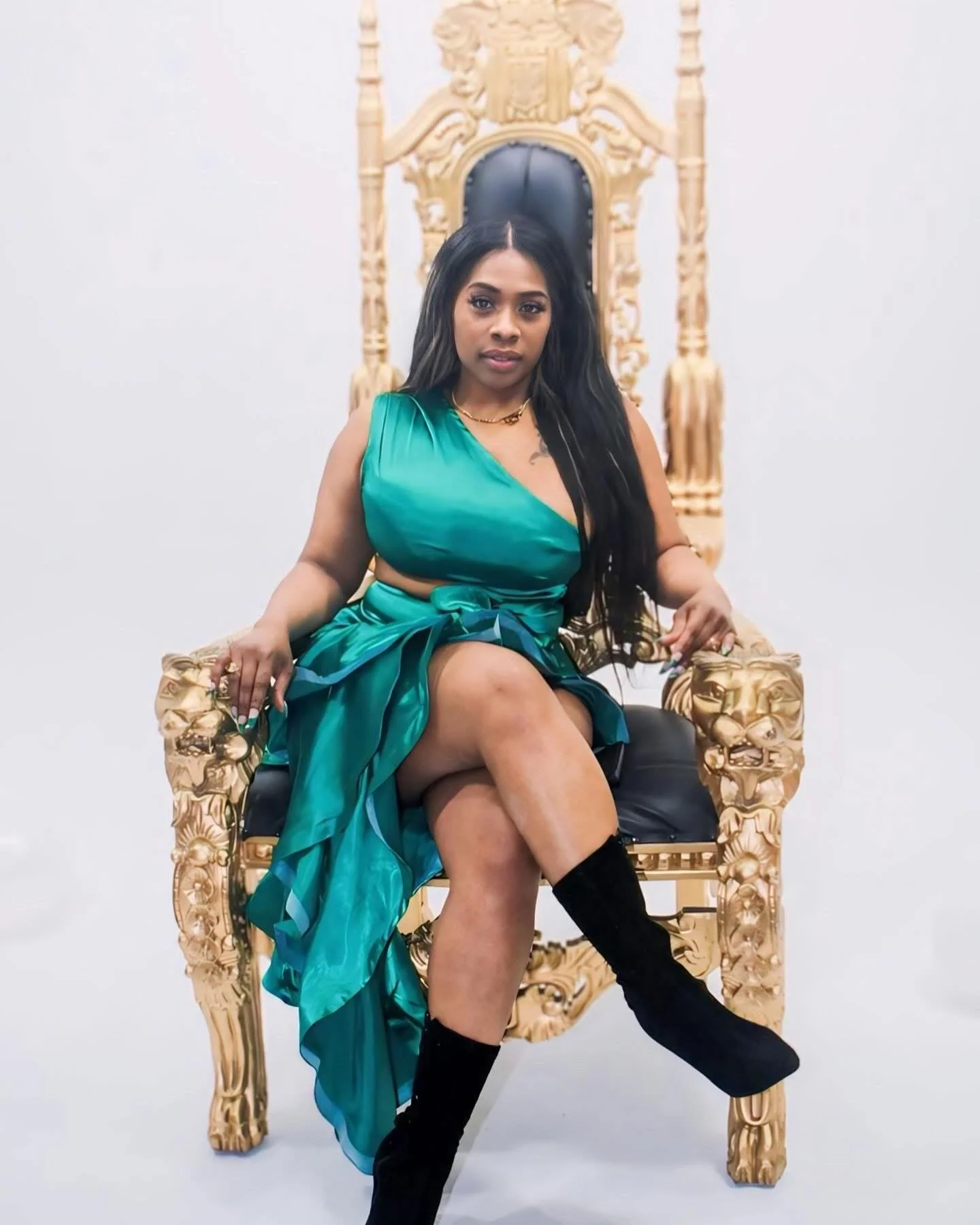 A woman sitting on an ornate, gold-colored throne with a black cushion, wearing a teal dress and black knee-high boots, against a plain white background.