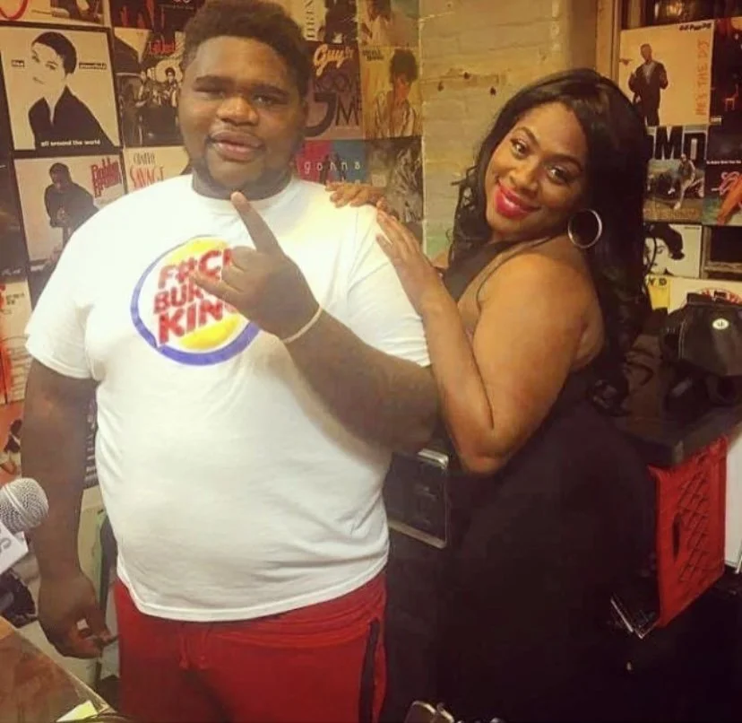 Two people, a man and a woman, posing together in a music or media room with posters on the wall. The man is wearing a white T-shirt with a Burger King logo, and the woman is dressed in black with large hoop earrings, smiling and touching the man's s