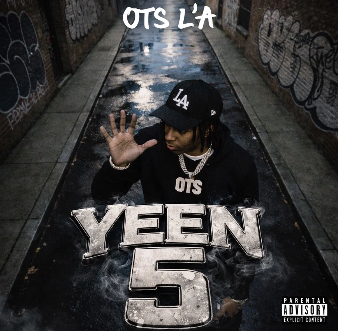 A young man wearing a black 'LA' baseball cap, black hoodie, and jewelry, crouched in a narrow, graffiti-lined alley with puddles on the ground. He is holding a large sign that says 'YEEZY 5' with smoke effects around it. The text 'OTS L'A' is at the