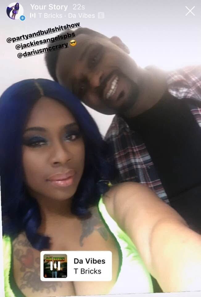 A woman with blue hair and visible tattoos taking a selfie with a smiling man behind her. Both are in casual clothing, and the woman has bright makeup.