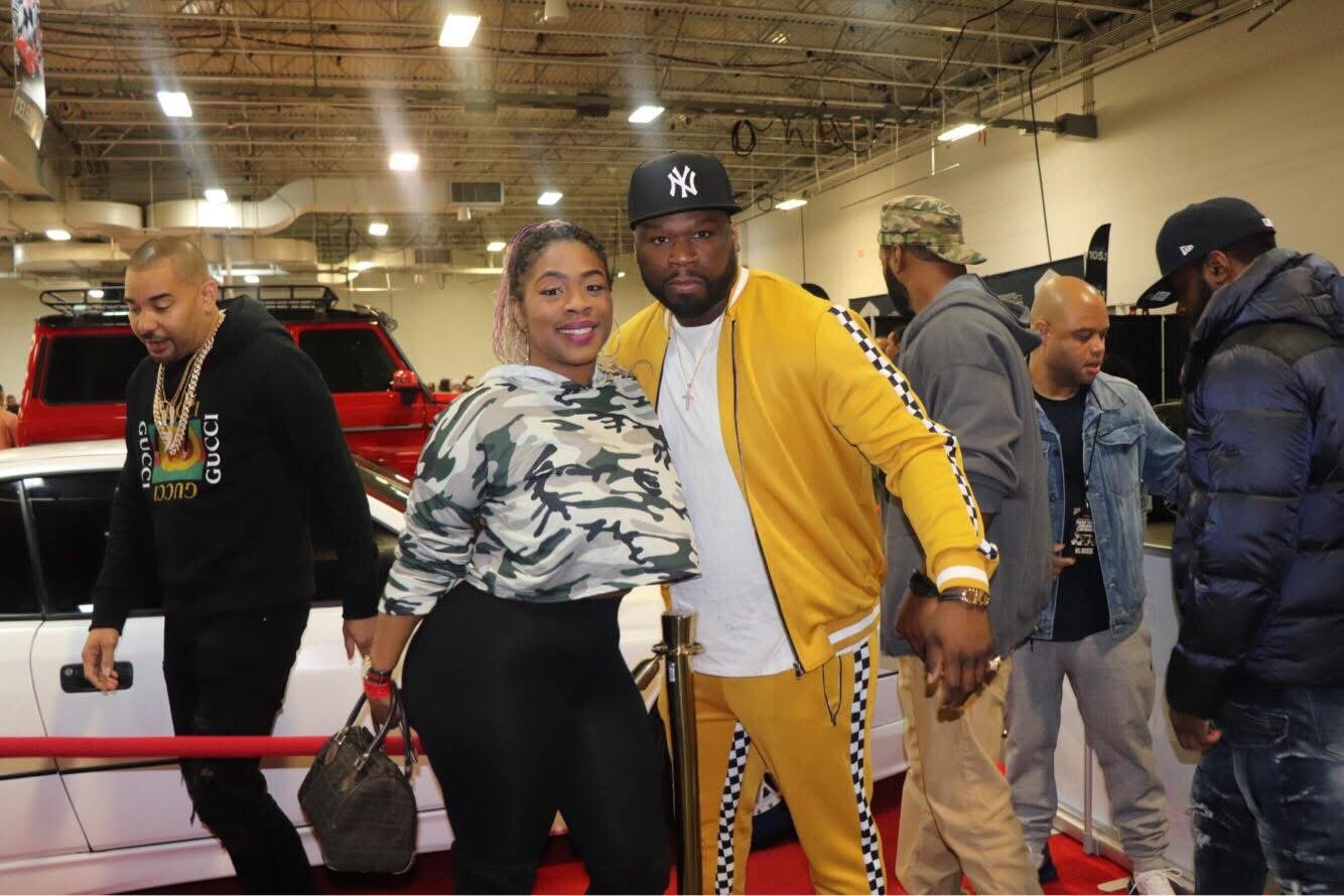 A group of people at an indoor car show. Two individuals in the foreground are posing for the camera; a woman in a camouflage hoodie and a man in a yellow tracksuit. Other attendees are looking at cars in the background.