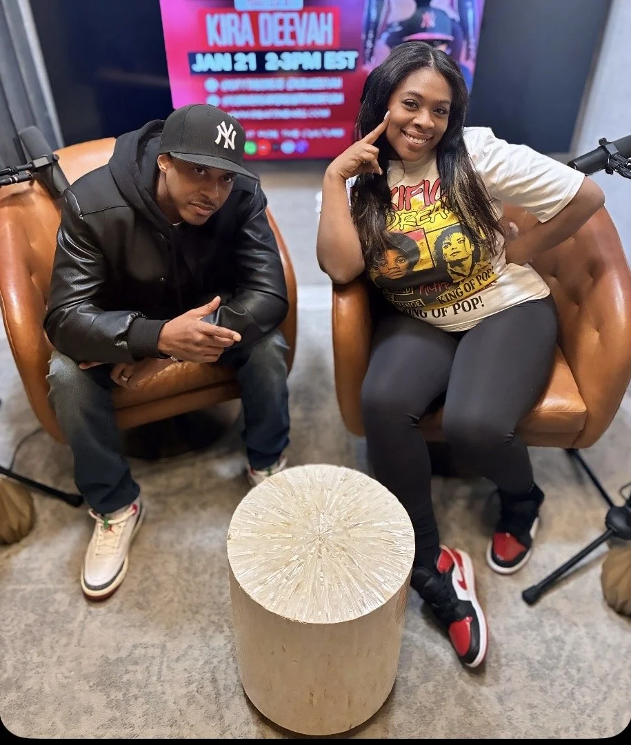 A woman and a man sitting on brown leather chairs in a studio setting with a round wooden table in front of them. The woman is smiling, wearing a graphic T-shirt and black leggings, while the man is looking serious, wearing a black jacket, grey pants