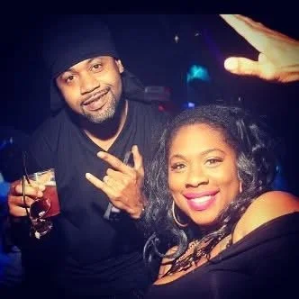 Man and woman smiling at the camera during a celebration, with the man holding a drink and making a peace sign, and the woman wearing hoop earrings and smiling broadly.