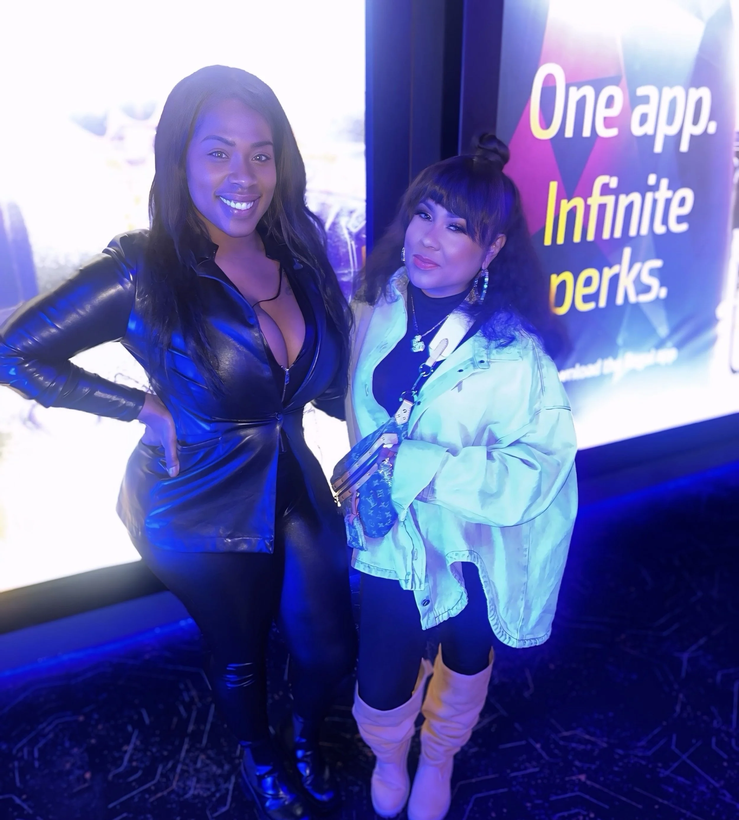 Two women standing together in front of a colorful illuminated sign with the text 'One app. Infinite perks.' The woman on the left is wearing a shiny black leather suit and smiling, while the woman on the right is dressed in a yellow jacket, black le
